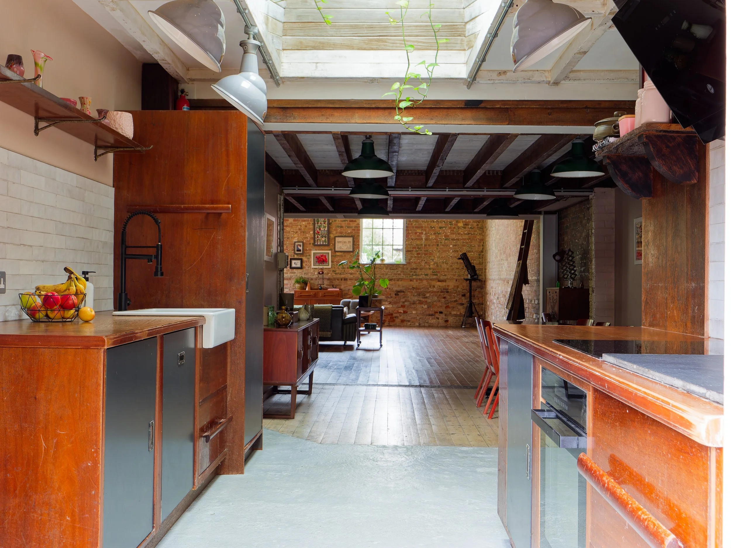 Open-concept kitchen and living room with exposed brick walls, wooden beams, and large windows, featuring a kitchen island, fruit basket, and seating area.