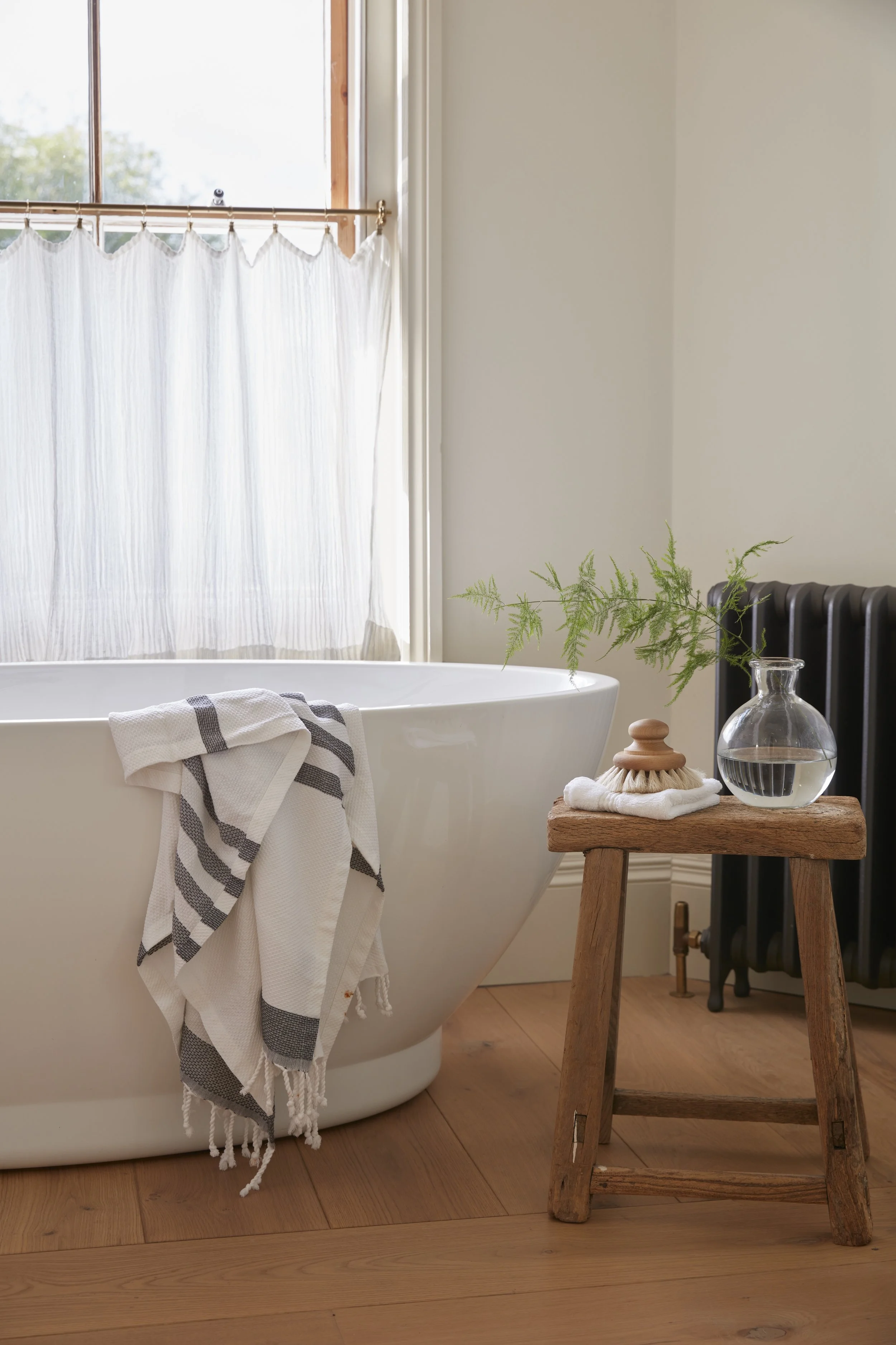 A cozy bathroom corner with a white bathtub, a towel hanging on its edge, a small wooden stool with a glass vase containing greenery, a folded towel, and a decorative brush, next to a black radiator and a window with white curtains.