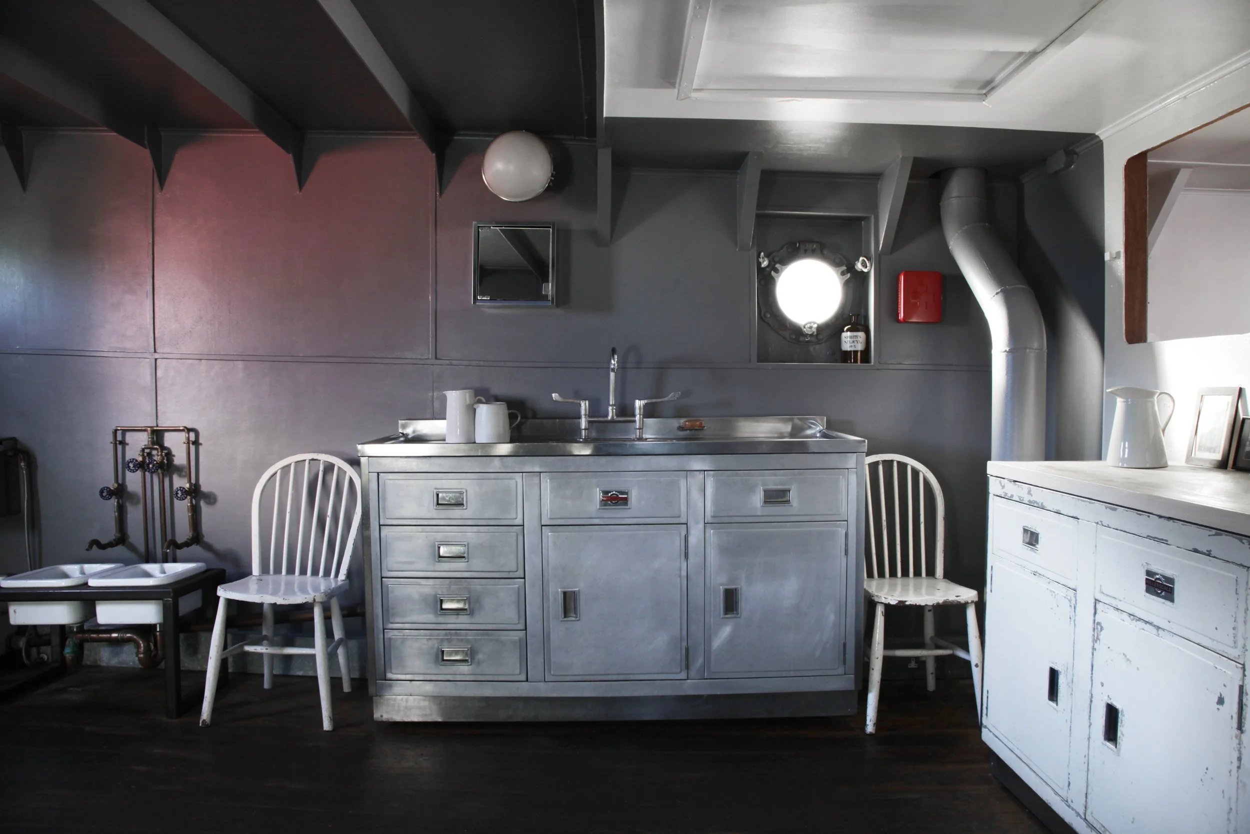 A rustic kitchen with dark and white cabinets, a metal sink, white chairs, a framed photo, jug, and small items on top. There are metal pipes, a window, and various kitchen fixtures.
