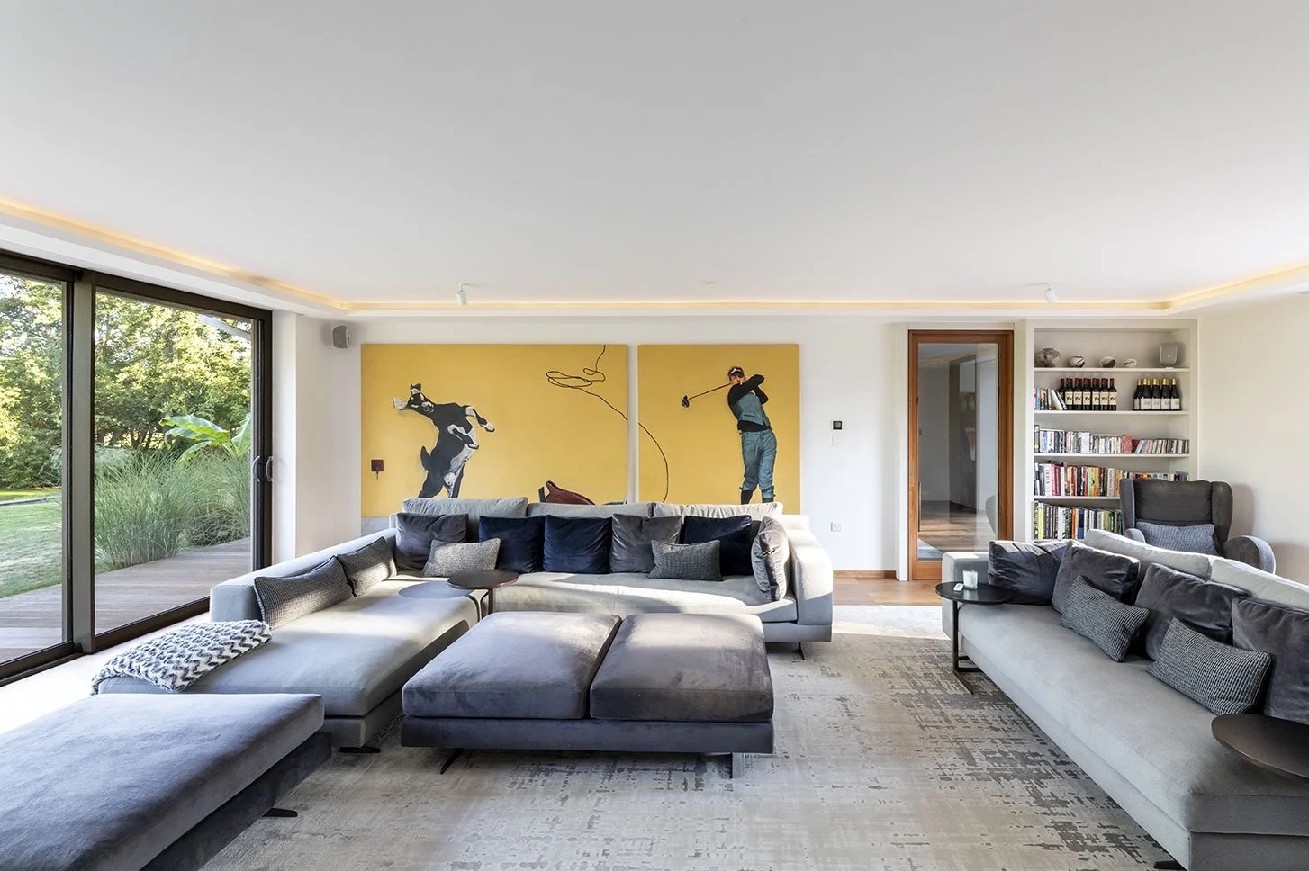 A spacious living room with large glass sliding doors overlooking a garden. The room features a sectional sofa with gray and blue cushions, a gray ottoman, a patterned throw blanket, a black armchair, and a bookshelf filled with books and decorative 