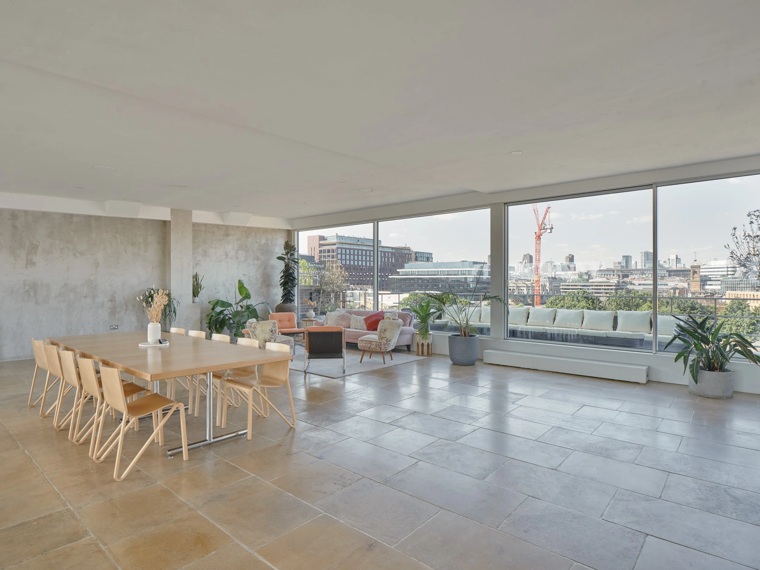 Modern open-plan living room and dining area with large windows overlooking cityscape, featuring a dining table with nine wooden chairs, a sitting area with sofas and armchairs, numerous potted plants, and natural light.