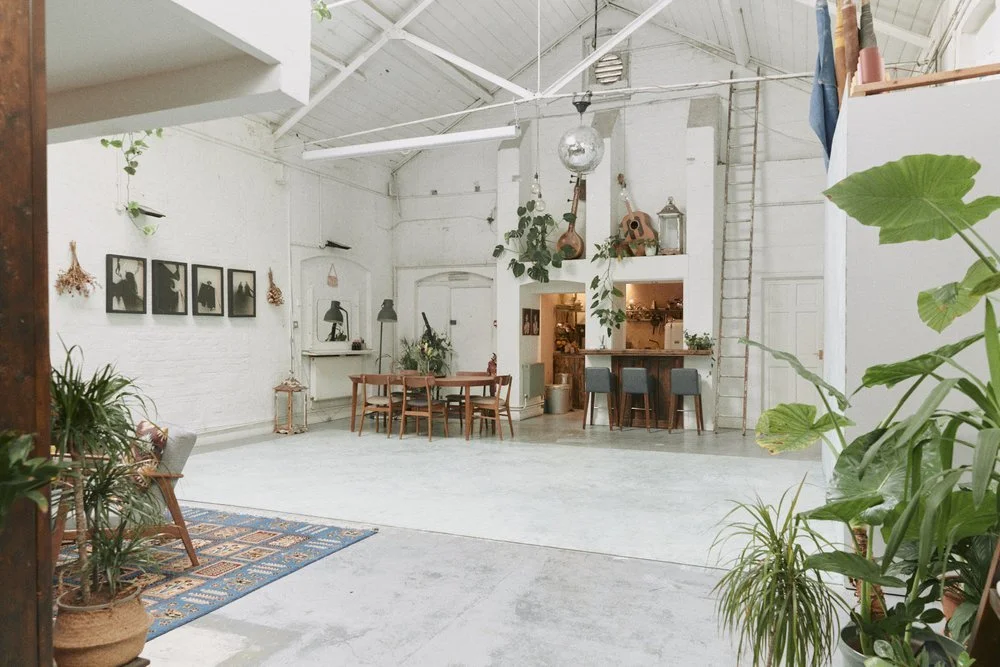 Spacious, modern interior with high vaulted ceiling, white walls, and large green potted plants. Features a dining area with a wooden table and six chairs, a bar counter with three blue stools, and decorative hanging art and musical instruments.