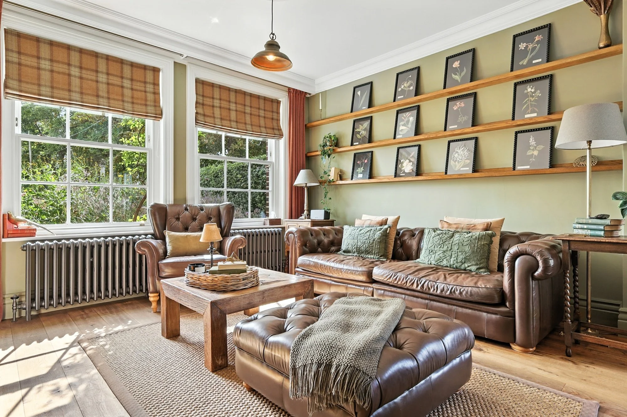 Cozy living room with large windows, plaid blinds, green walls, wooden shelves with framed flower artwork, leather sofa and armchair, wooden coffee table, and floor lamp.