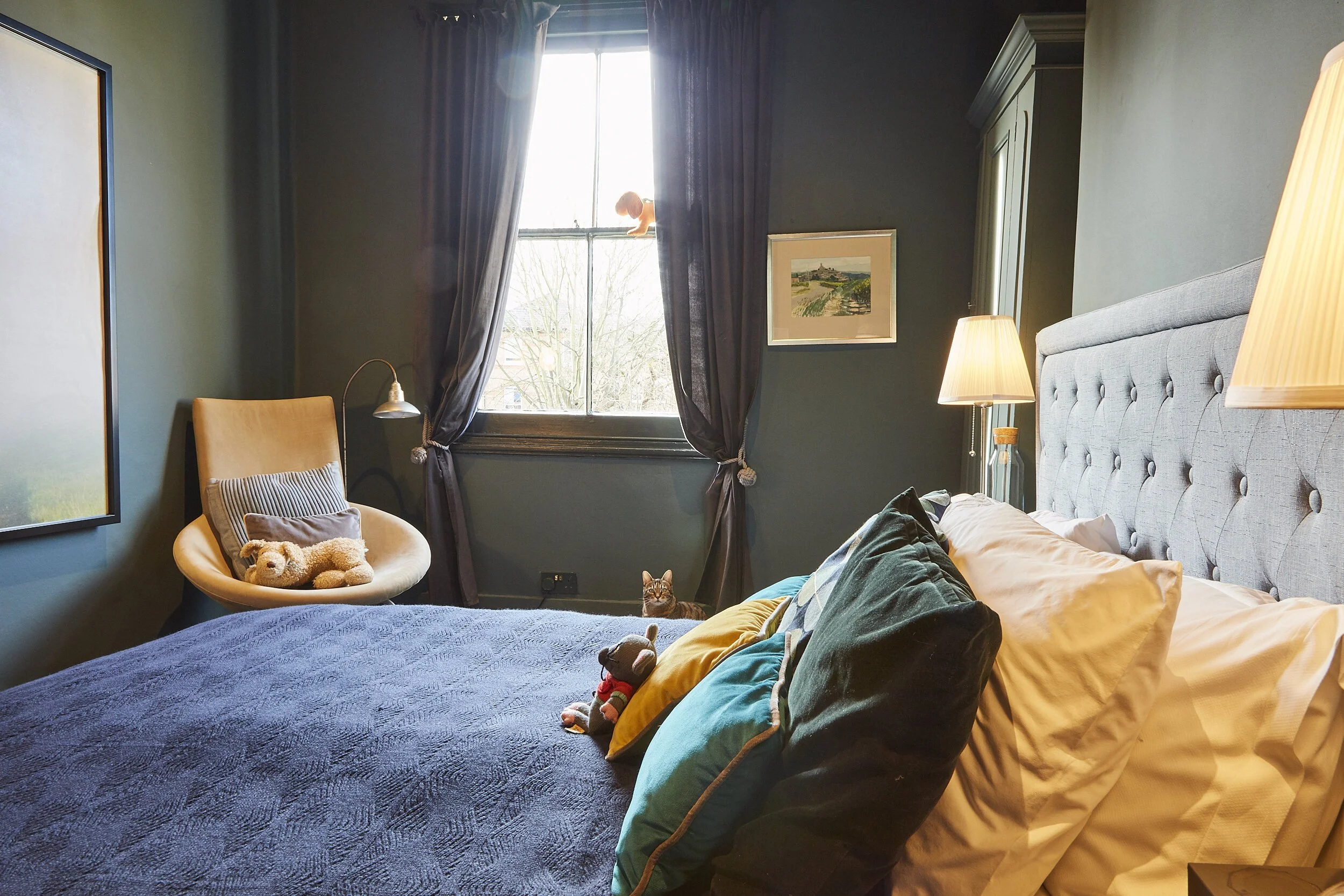 Cozy bedroom with a large bed covered in a blue quilt and multiple pillows, a chair with a teddy bear and a pillow, a window with dark curtains, a cat on the windowsill, framed artwork on the wall, and bedside lamps providing warm lighting.