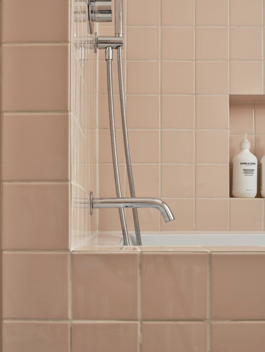Close-up of a pink-tiled shower with a chrome faucet, a handheld showerhead with flexible hoses, and a small shelf with a white bottle of shampoo or conditioner.