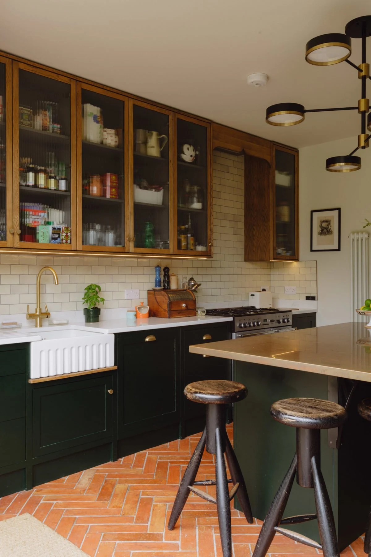 Kitchen with green lower cabinets, white farmhouse sink, brick floor, open wooden cabinets with glass doors, small plants, and stools at a breakfast bar.