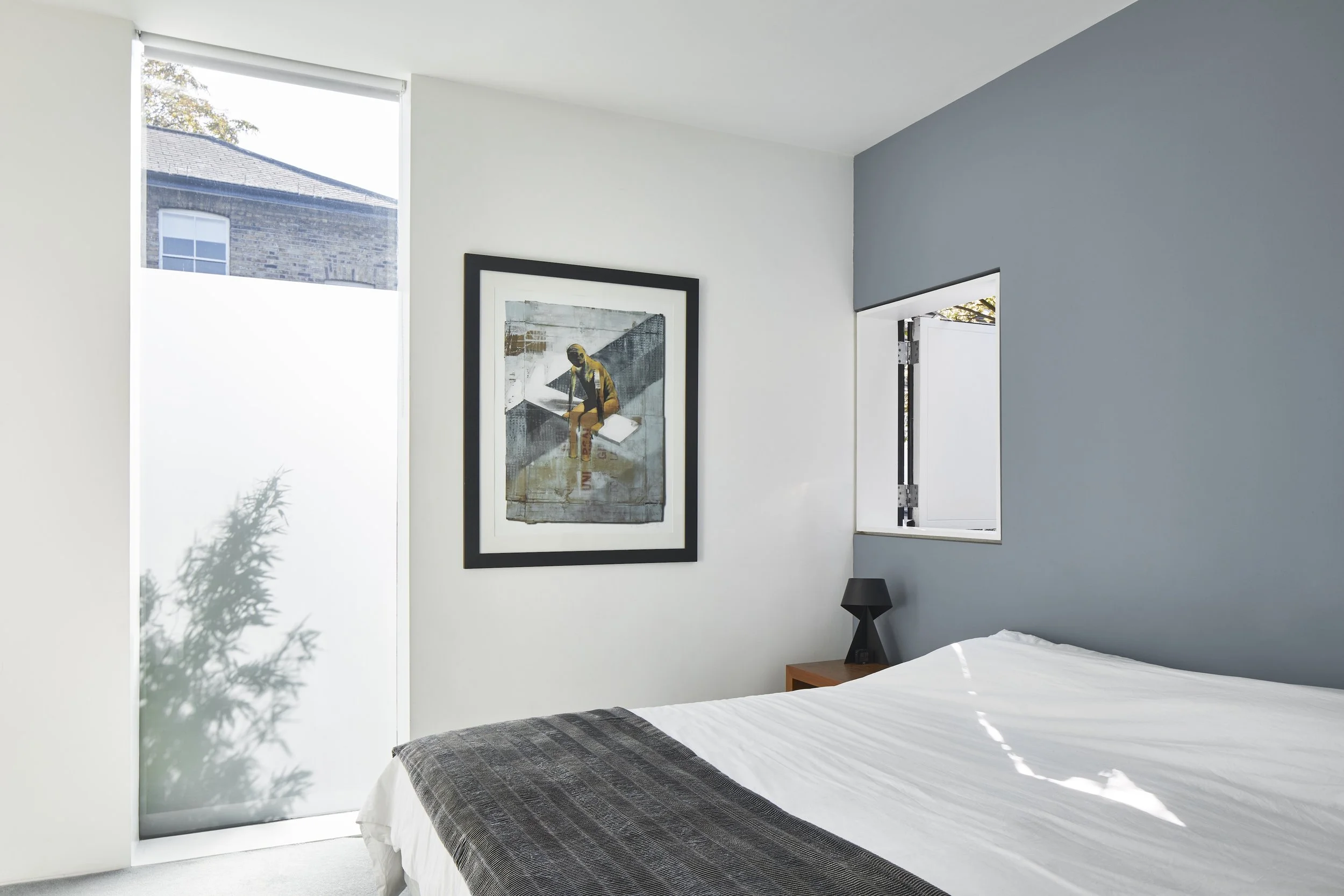Bedroom with white walls, a black-framed art piece, a small wooden nightstand with a black lamp, a large window, and a bed with white bedding and a dark gray blanket.