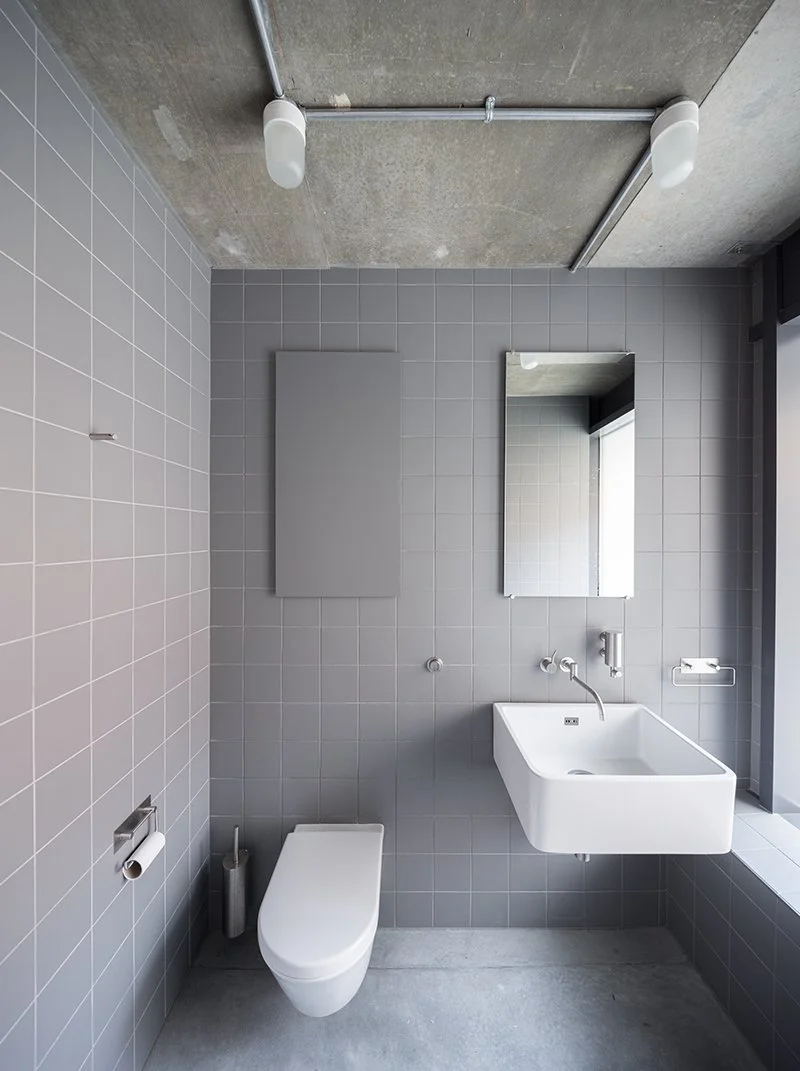Modern bathroom with gray tiled walls, wall-mounted white toilet, rectangular sink with a wall-mounted faucet, mirror, and minimal fixtures. Ceiling has exposed concrete with industrial-style lighting and piping.