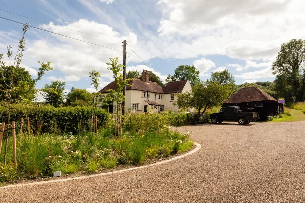 A country scene with a gravel driveway, a white house with a brown roof, a black truck, and a detached barn or garage, surrounded by green trees and plants under a partly cloudy sky.