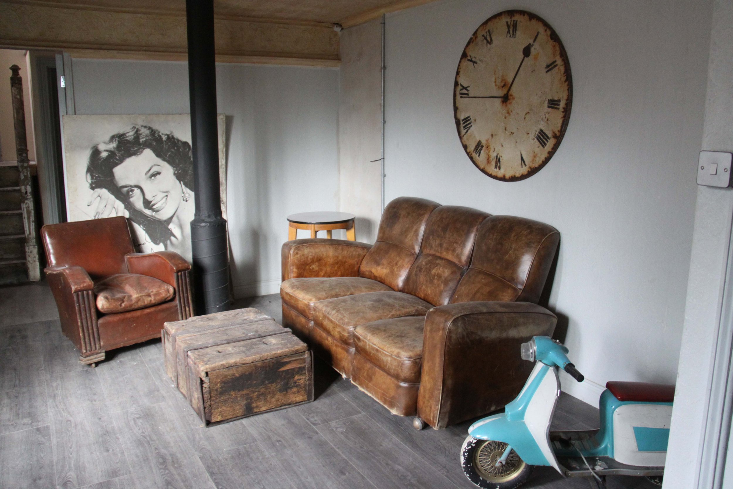 A vintage-style living room corner with a brown leather sofa, an old wooden chest as a table, a smaller brown armchair, a side table, a large rustic clock on the wall, a teal and white scooter, and a black-and-white portrait of a smiling woman.