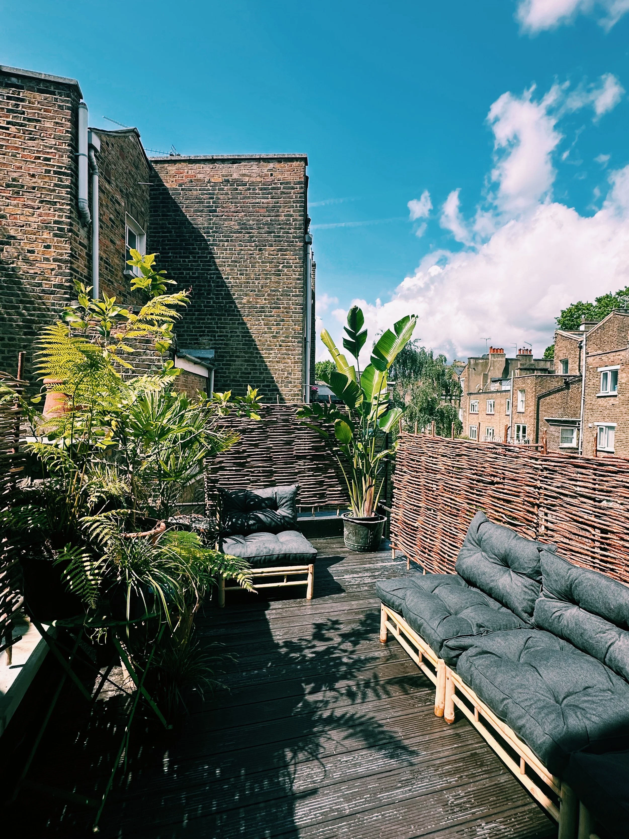 A rooftop terrace with couches, potted plants, and a woven fence under a bright blue sky with clouds.