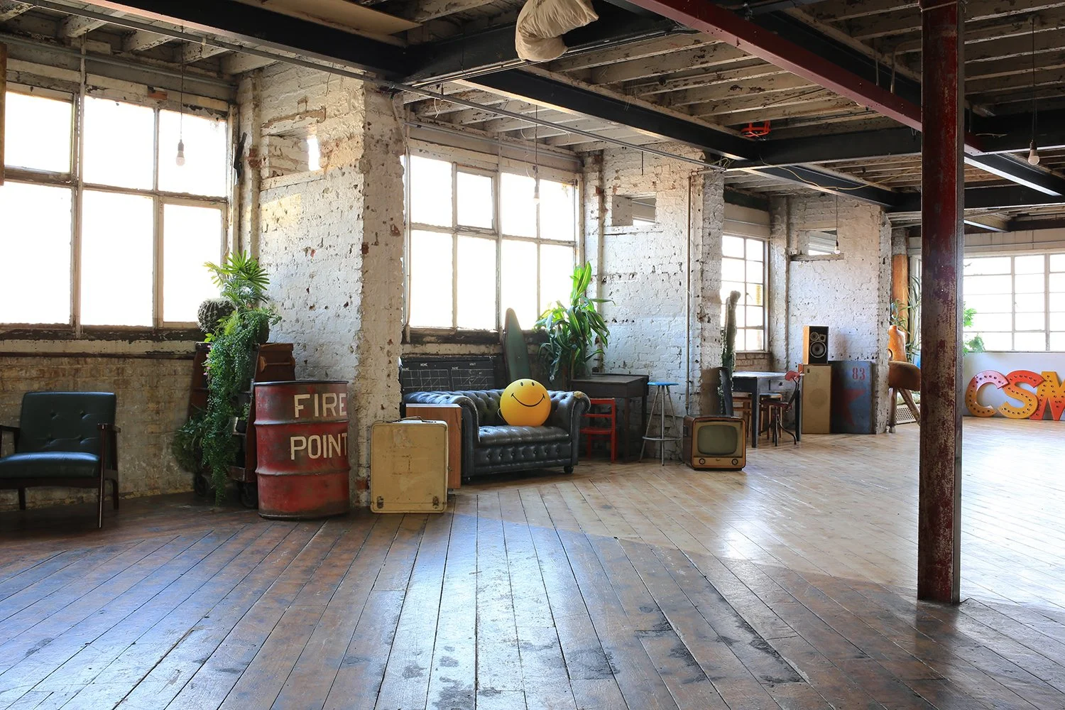 An industrial-style loft with exposed brick walls and large windows. The space features vintage furniture, plants, and decorative items like a smiley face ball, a small vintage television, and large colorful letters at the far end.