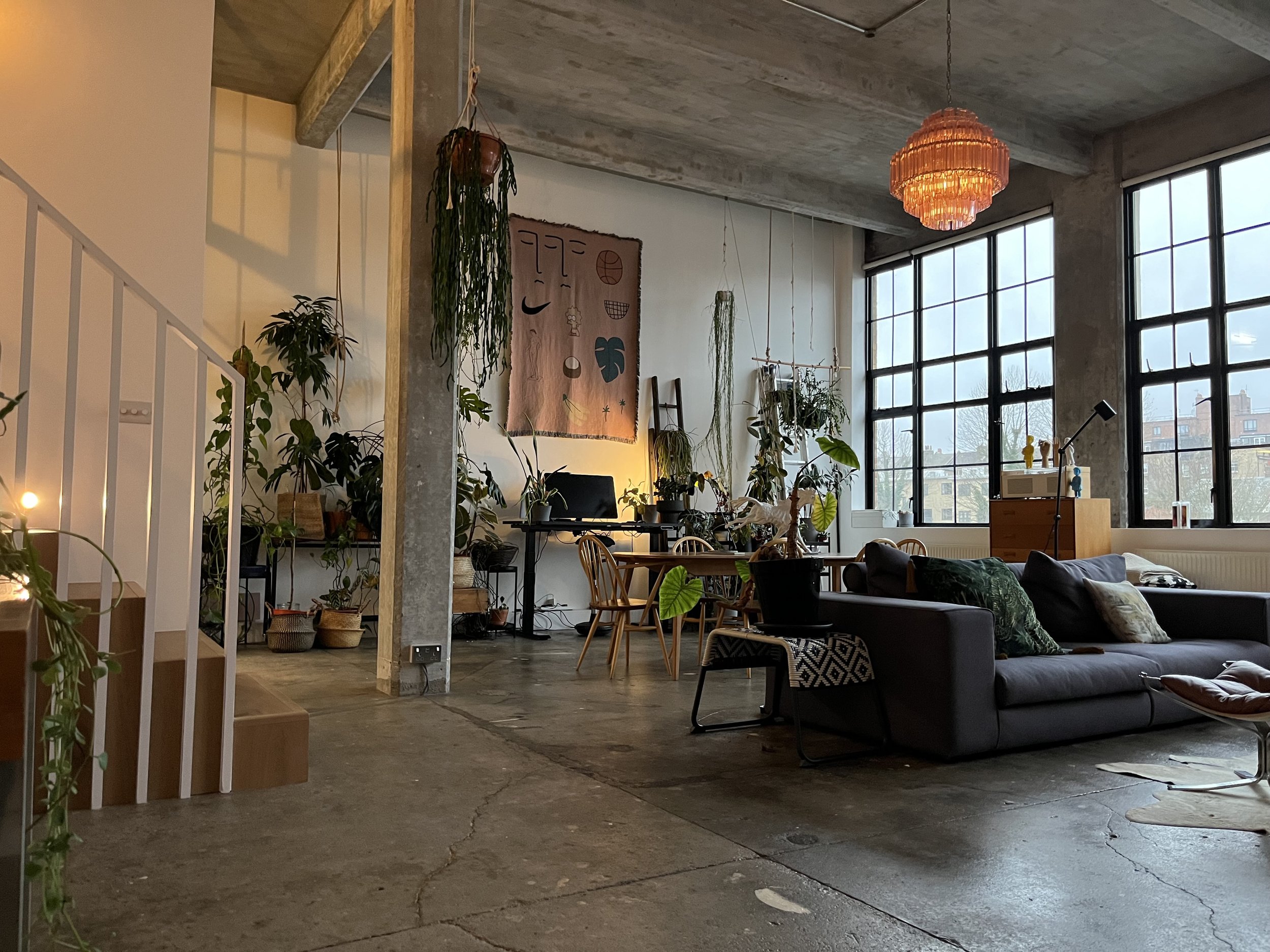 A spacious, industrial-style living room with large windows, concrete floors, and various potted plants. There is a dark sofa with cushions, a wooden side table, a computer desk with a monitor, and wall hanging art. A hanging pink geometric chandelie