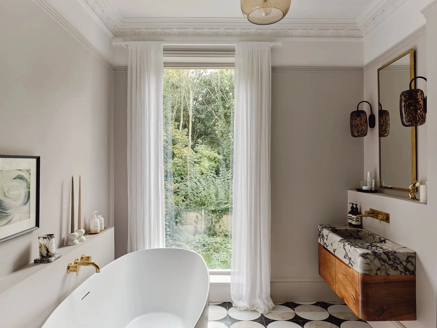 A luxurious bathroom with a large window overlooking greenery, a modern bathtub with gold fixtures, a marble sink vanity, a mirror, and elegant wall-mounted light fixtures.