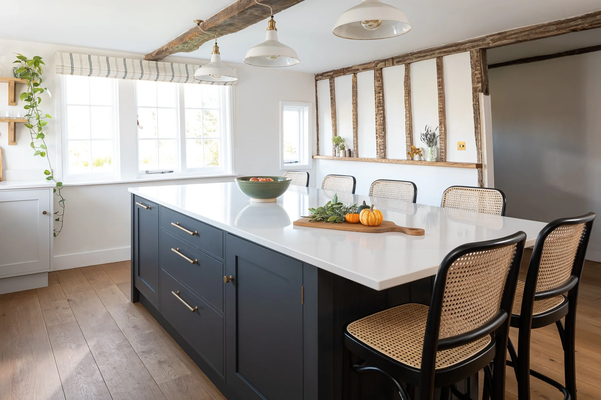 A modern kitchen with a white island counter, blue cabinets, and black chairs with rattan seats. Large windows with a striped valance, wooden ceiling beams, and hanging white pendant lights. Decor includes a fruit bowl, pumpkins, and plants on shelve
