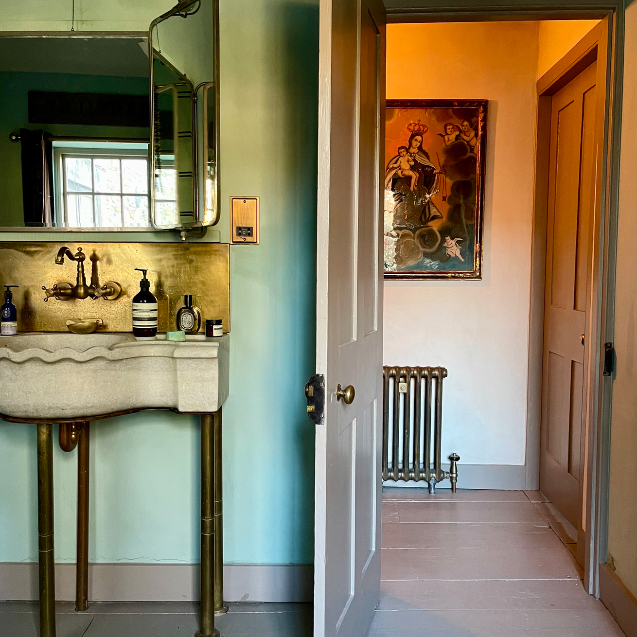 A vintage bathroom with a cream-colored sink and gold fixtures, seen through an open door leading to a hallway with a radiator, artwork on the wall, and a wooden door to another room.