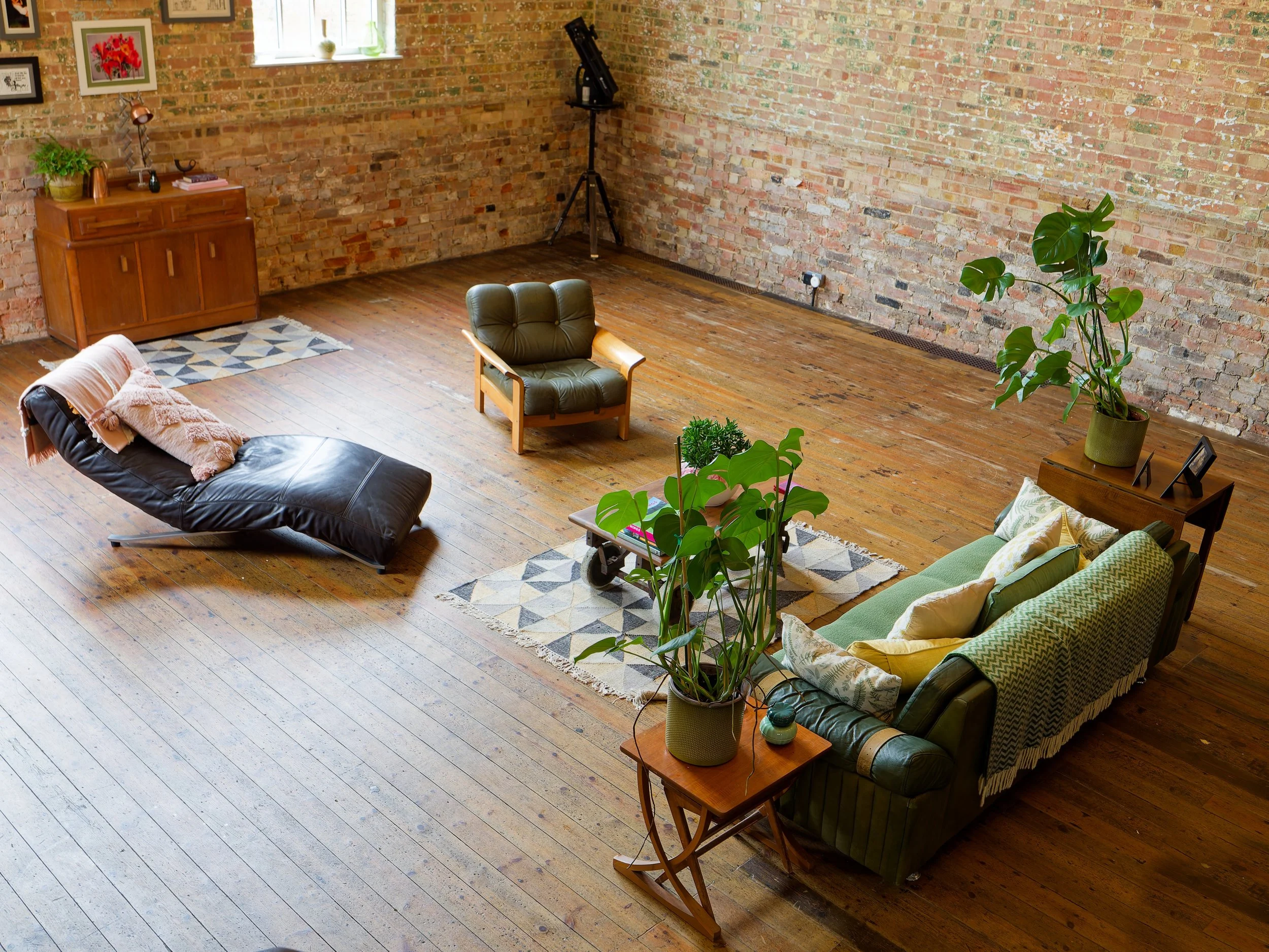 Spacious living room with wooden floors and exposed brick walls, furnished with a green couch, a black leather lounge chair, a green upholstered armchair, potted plants, a wooden sideboard, and a small wooden end table. There are framed pictures on t