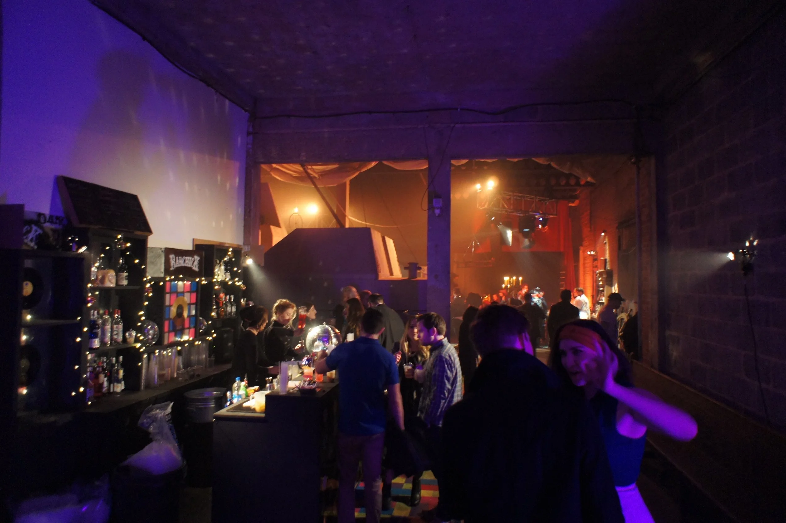 People socializing and dancing in a dark nightclub or concert venue with blue and orange lighting, exposed brick walls, and a bar area.