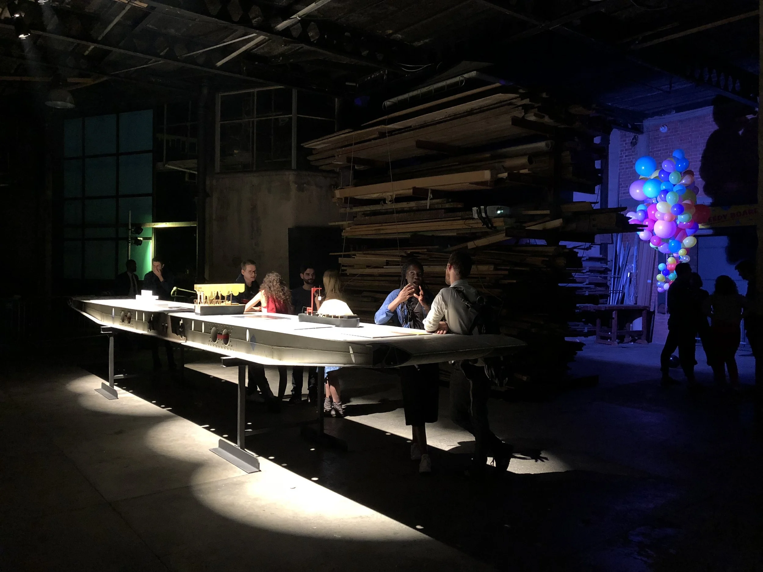 People socializing and drinking at a bar in a dimly lit industrial-style venue with a large pile of wooden planks as part of the decor and a cluster of colorful balloons in the background.