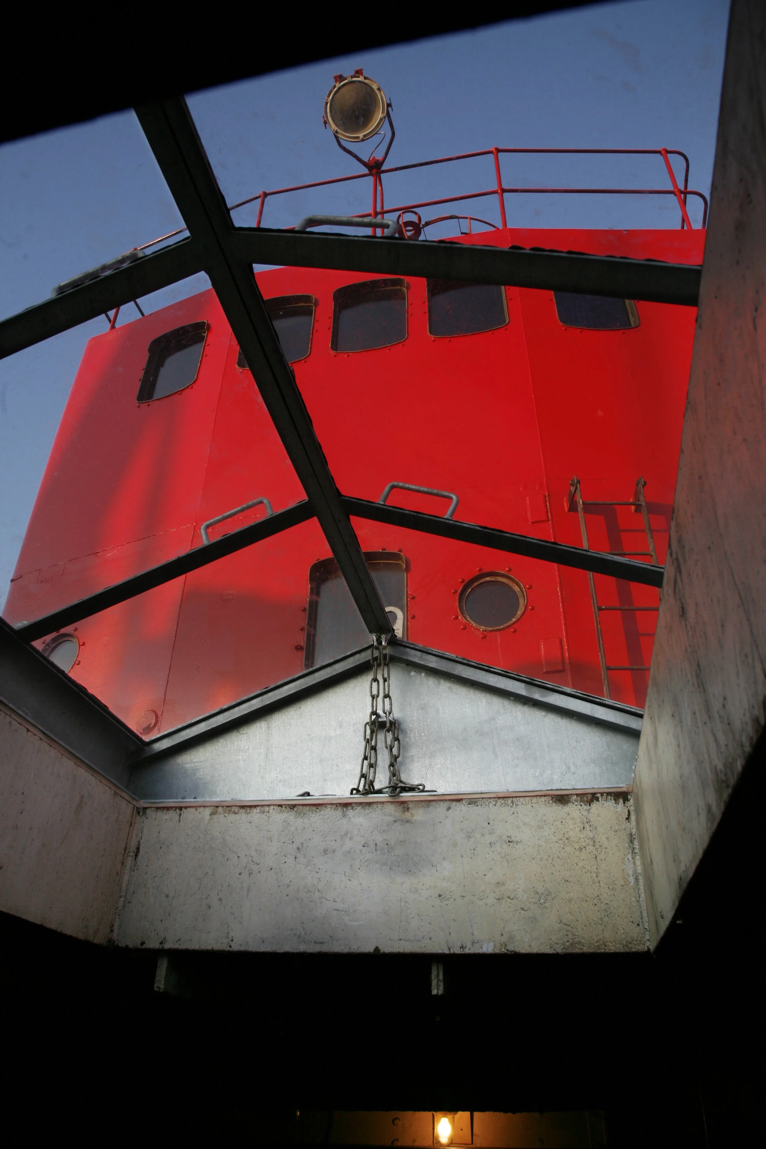 View of a red lighthouse viewed through a square window with black metal bars, chain, and a metal frame at the bottom, under a clear blue sky.