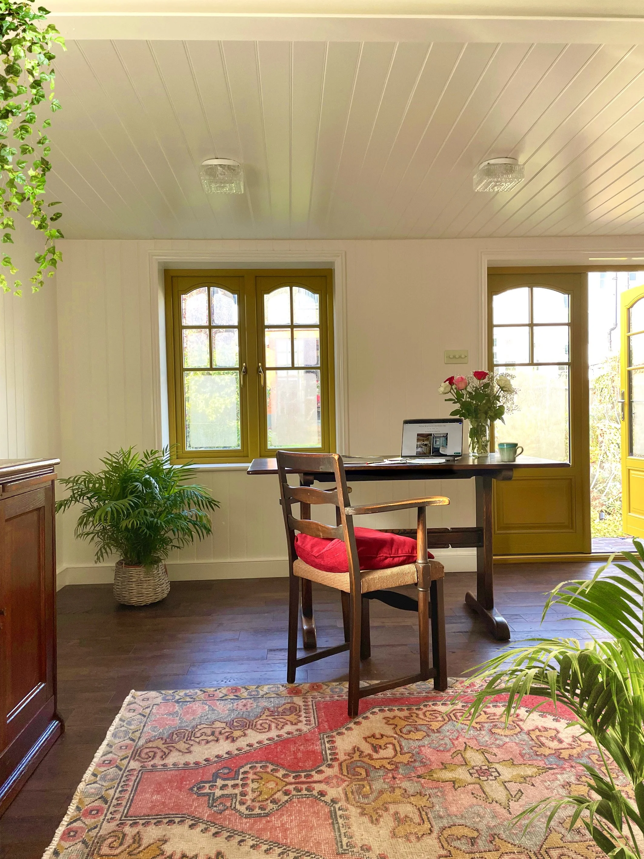 A cozy home office with a wooden desk, a chair with a red cushion, potted plants, a laptop, a vase of flowers, and a colorful patterned rug, with windows and a door letting in natural light.