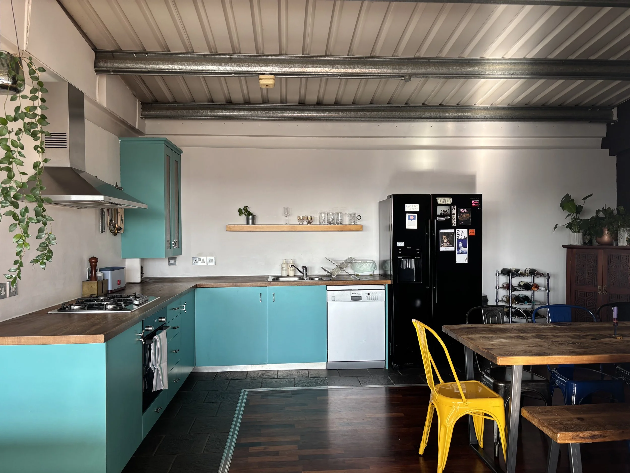 A kitchen with turquoise cabinets, a washer, a black refrigerator, a wooden table with yellow and black chairs, and plants.