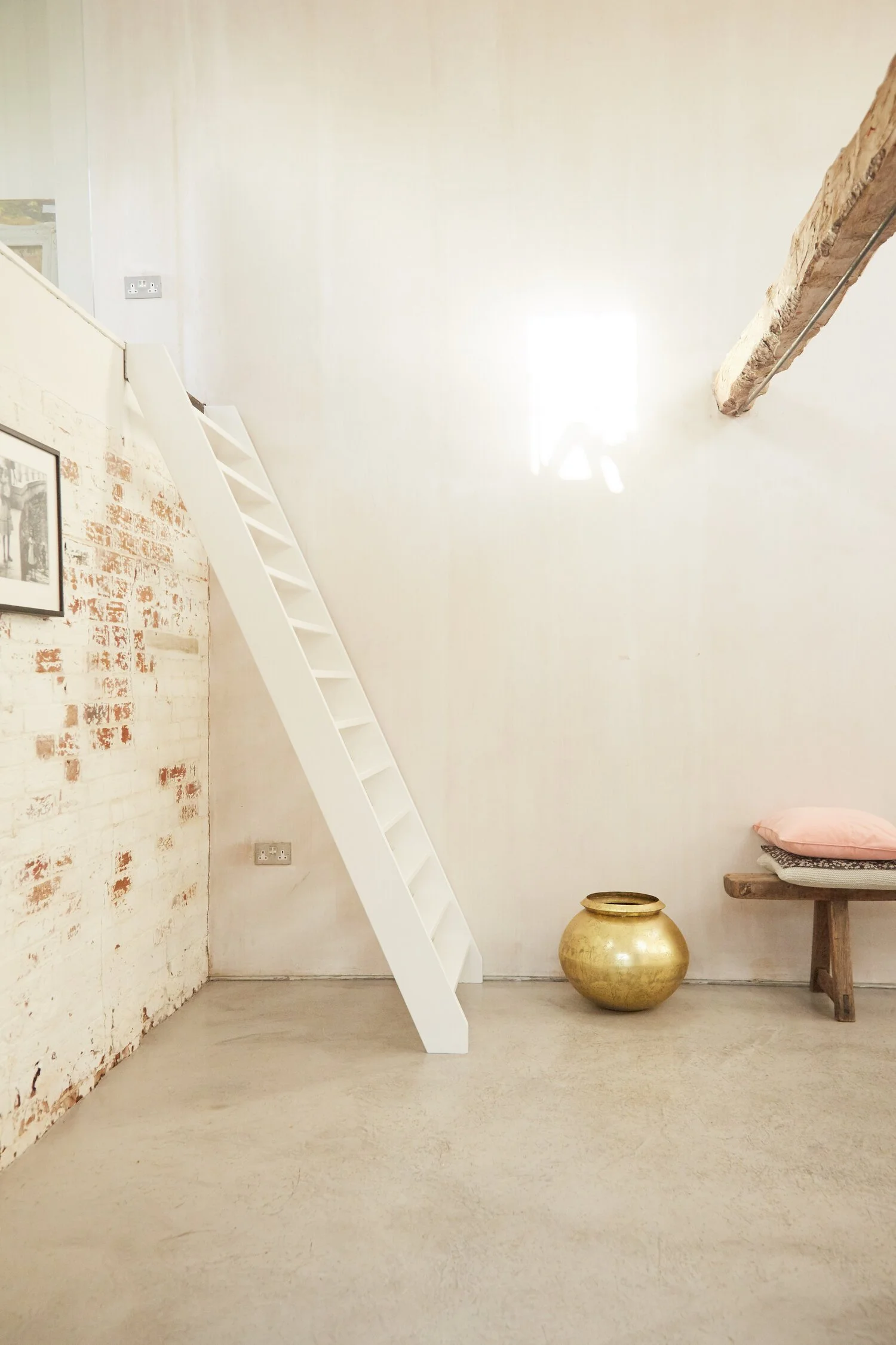 Minimalist interior with a white ladder leading to a loft, a gold decorative pot on the floor, and a wooden bench with pillows.