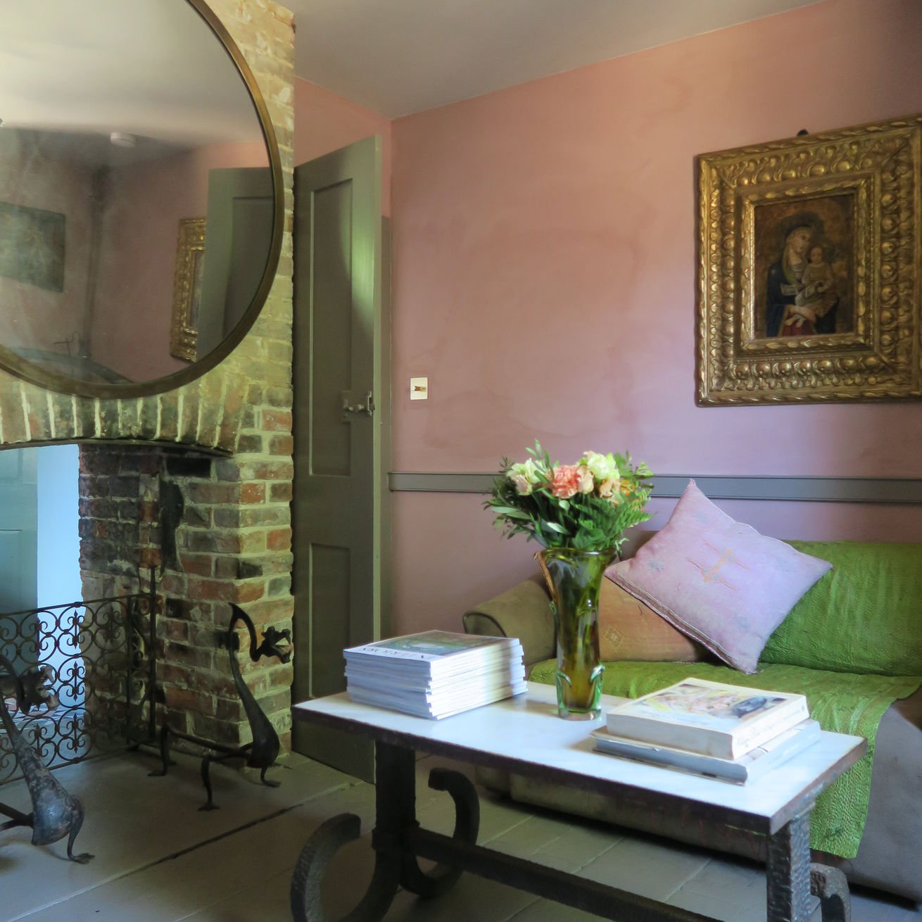 A cozy living room corner with a green sofa adorned with pink cushions, a white coffee table holding a vase with pink and white flowers and a stack of magazines or books, a pink wall with a framed religious painting, a brick fireplace with a large ro