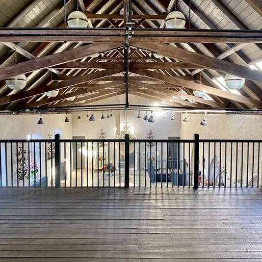 View of an indoor space with a high wooden ceiling, exposed beams, and a balcony with a wooden floor and black railing, overlooking a sunlit area with decorations and seating.