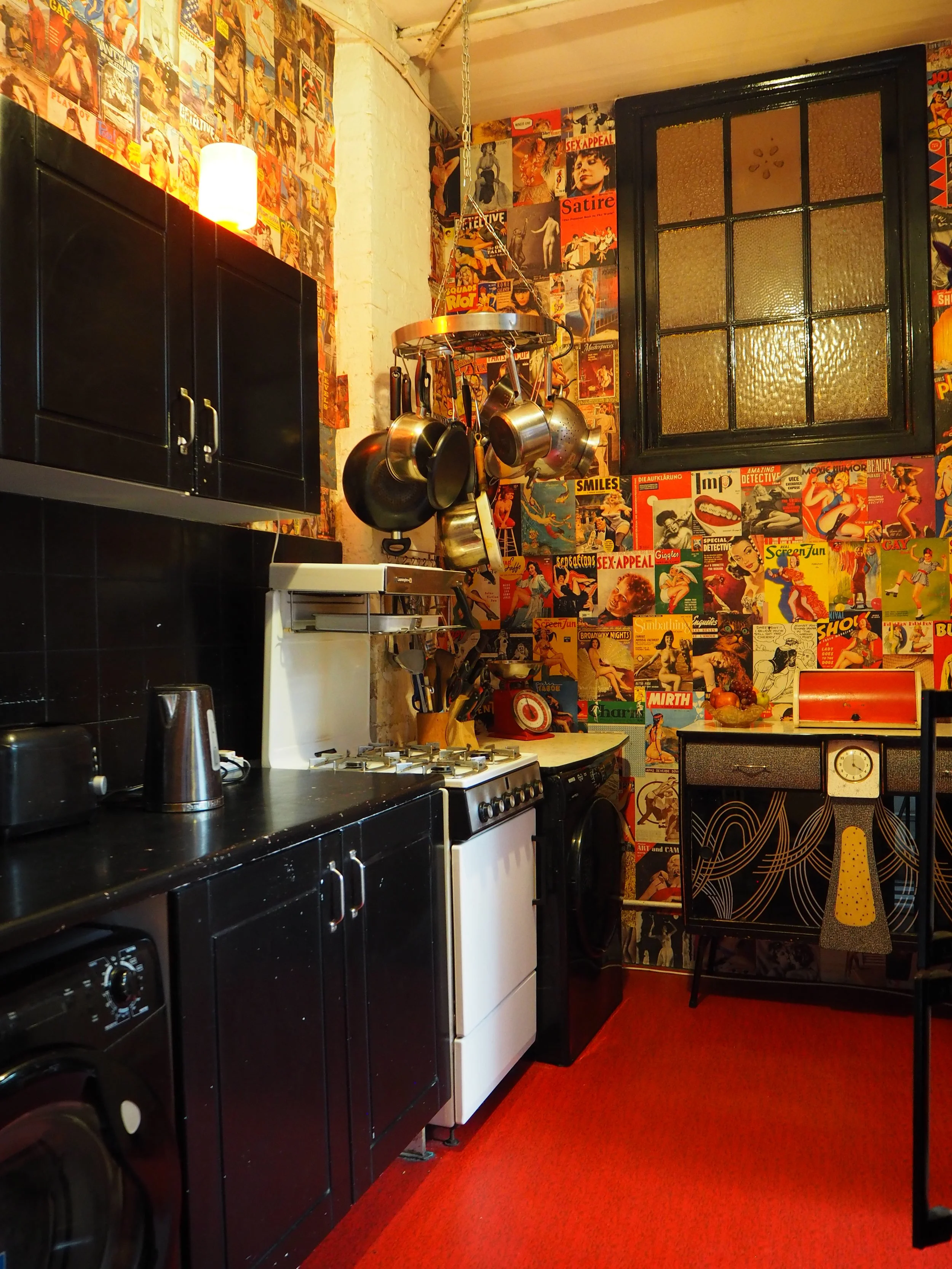 A kitchen with colorful vintage magazine cover wallpaper, black cabinets, a white stove, kitchen utensils hanging from a pot rack, and red carpet flooring.
