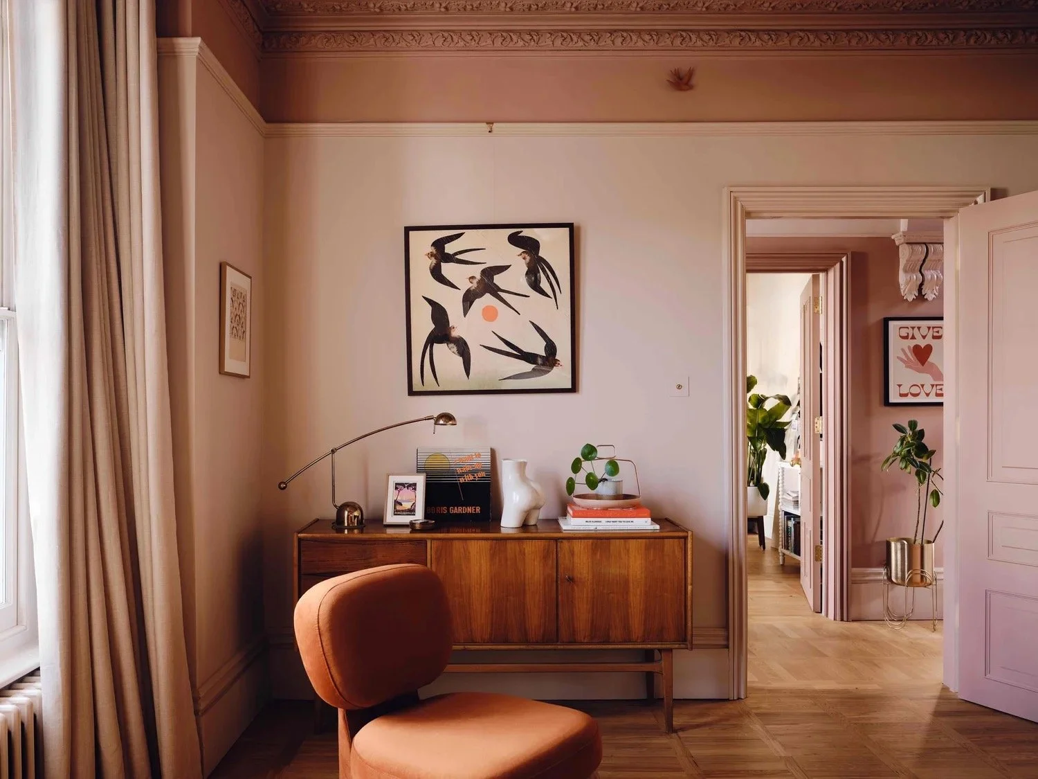 Elegant living room corner with a wooden sideboard decorated with books, plants, and ornaments, a vintage-style chair, framed artwork on the walls, beige curtains, and a view into an adjoining room with more plants and artwork.