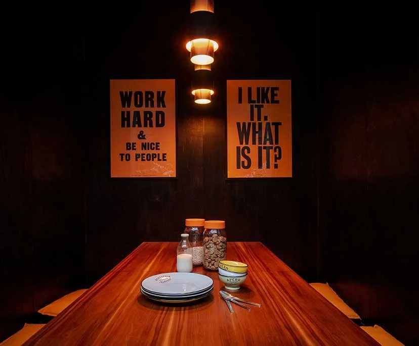 A wooden dining table with plates, spoon, cereal jars, milk, and butter container in a dark cozy room. Two motivational posters hang on the wall, one saying "Work hard & be nice to people" and the other asking "I like it. What is it?" Underneath the 