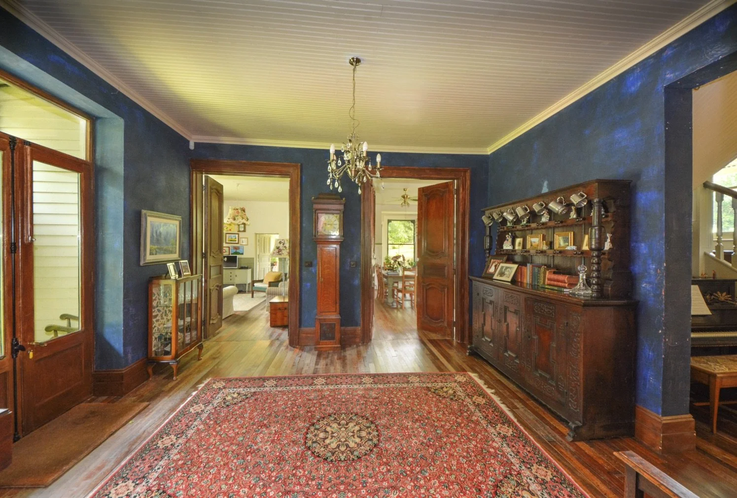 Interior of a vintage home with hardwood floors, dark blue walls, wood trim, a red decorative rug, a chandelier, and antique furniture including a cabinet, a grandfather clock, and framed photos.