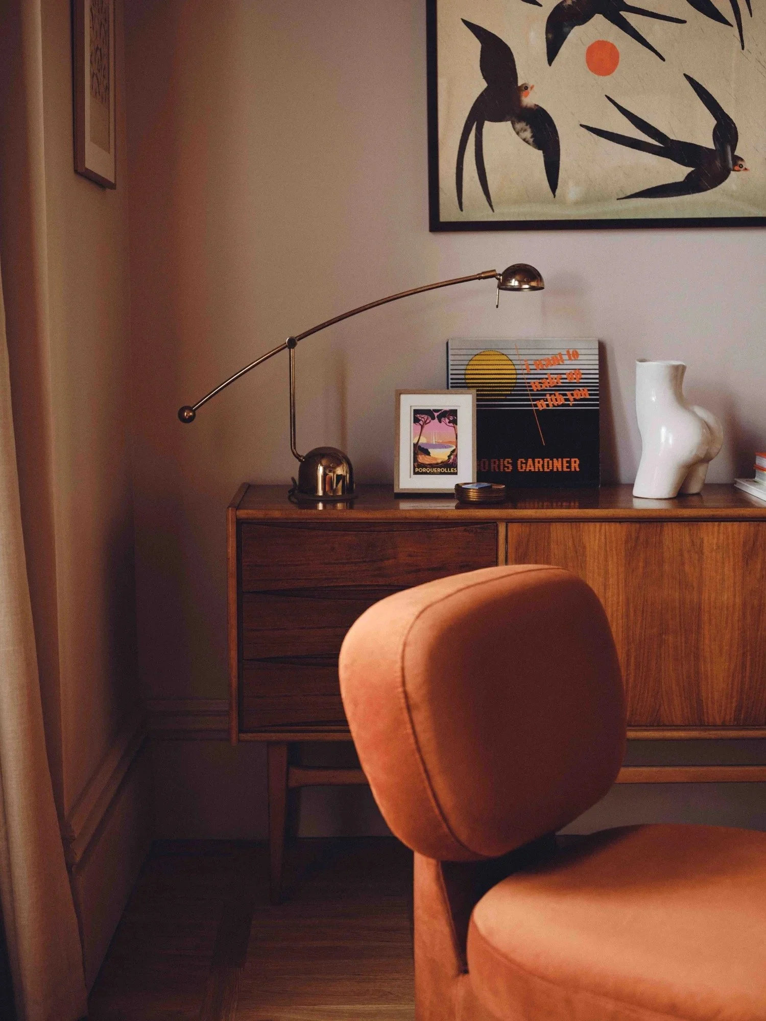 A cozy room corner with a wooden sideboard, a pink upholstered chair, gallery wall art, and a desk lamp on the sideboard.