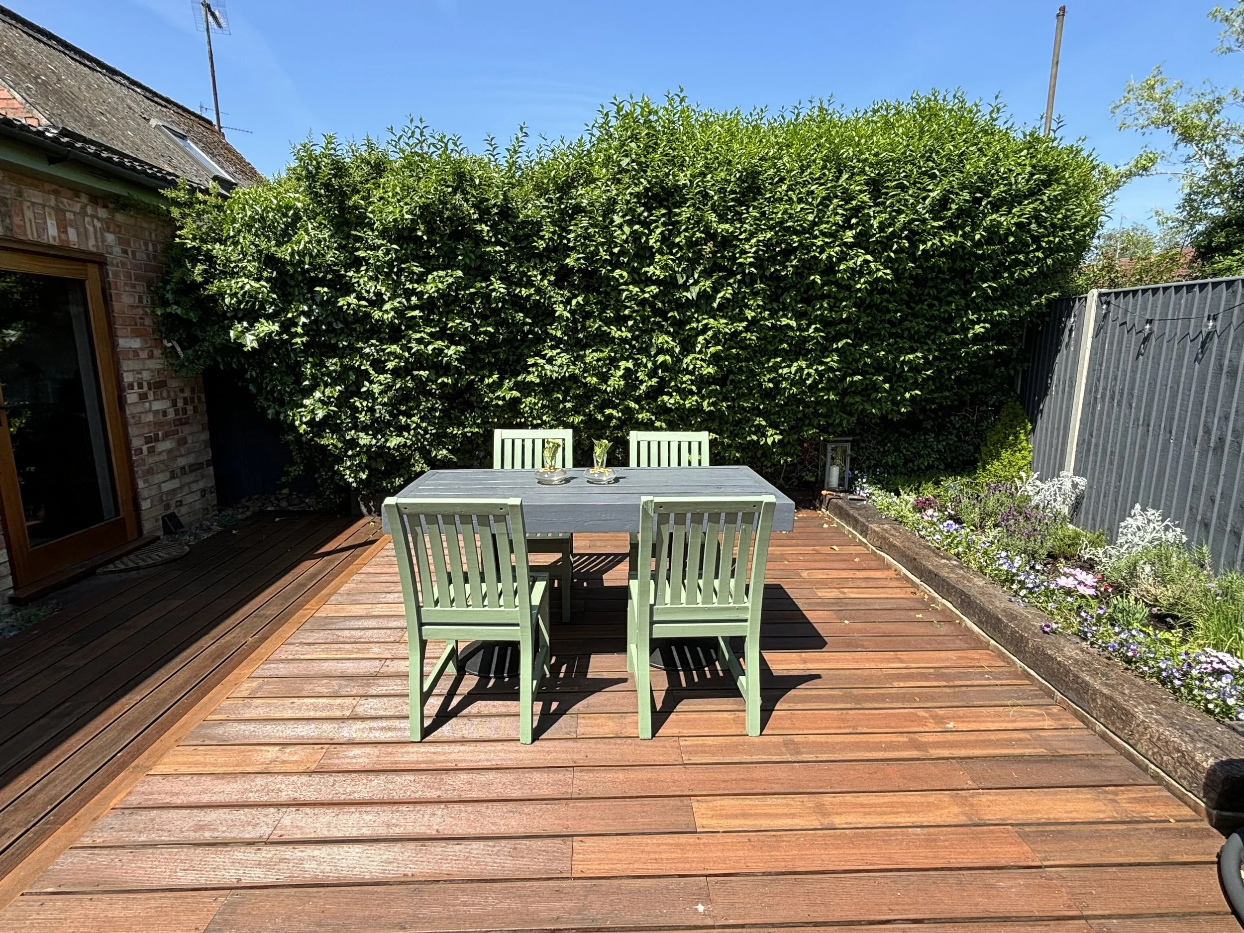 An outdoor wooden deck with a rectangular table and four green chairs, surrounded by greenery and a flower bed under a clear blue sky.