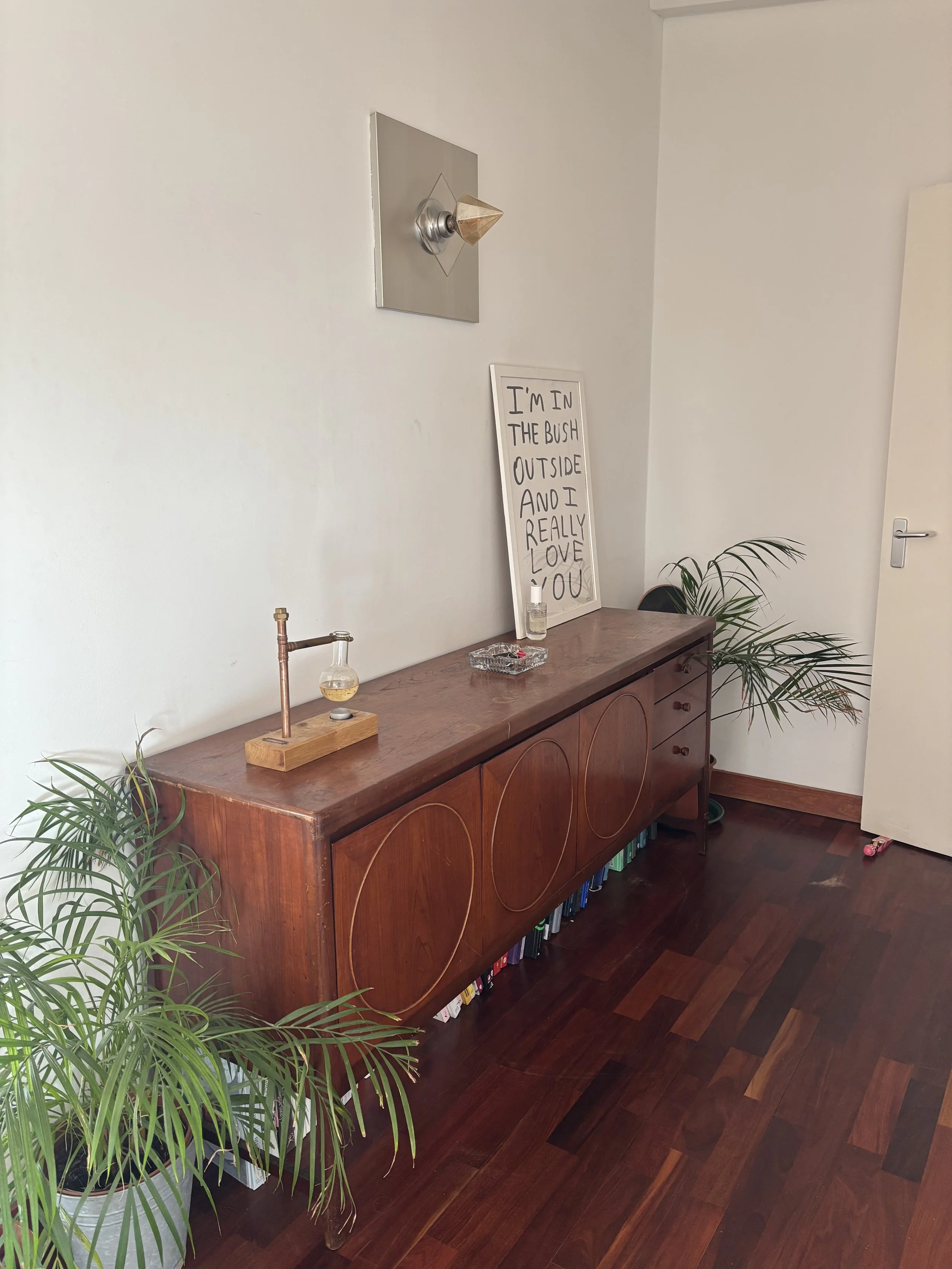 A wooden sideboard with circular patterns on its front, positioned against a white wall. On top of the sideboard are a small glass bottle with a liquid, an ashtray, and a white sign with handwritten text that reads "I'M IN THE BUSH OUTSIDE AND I REAL