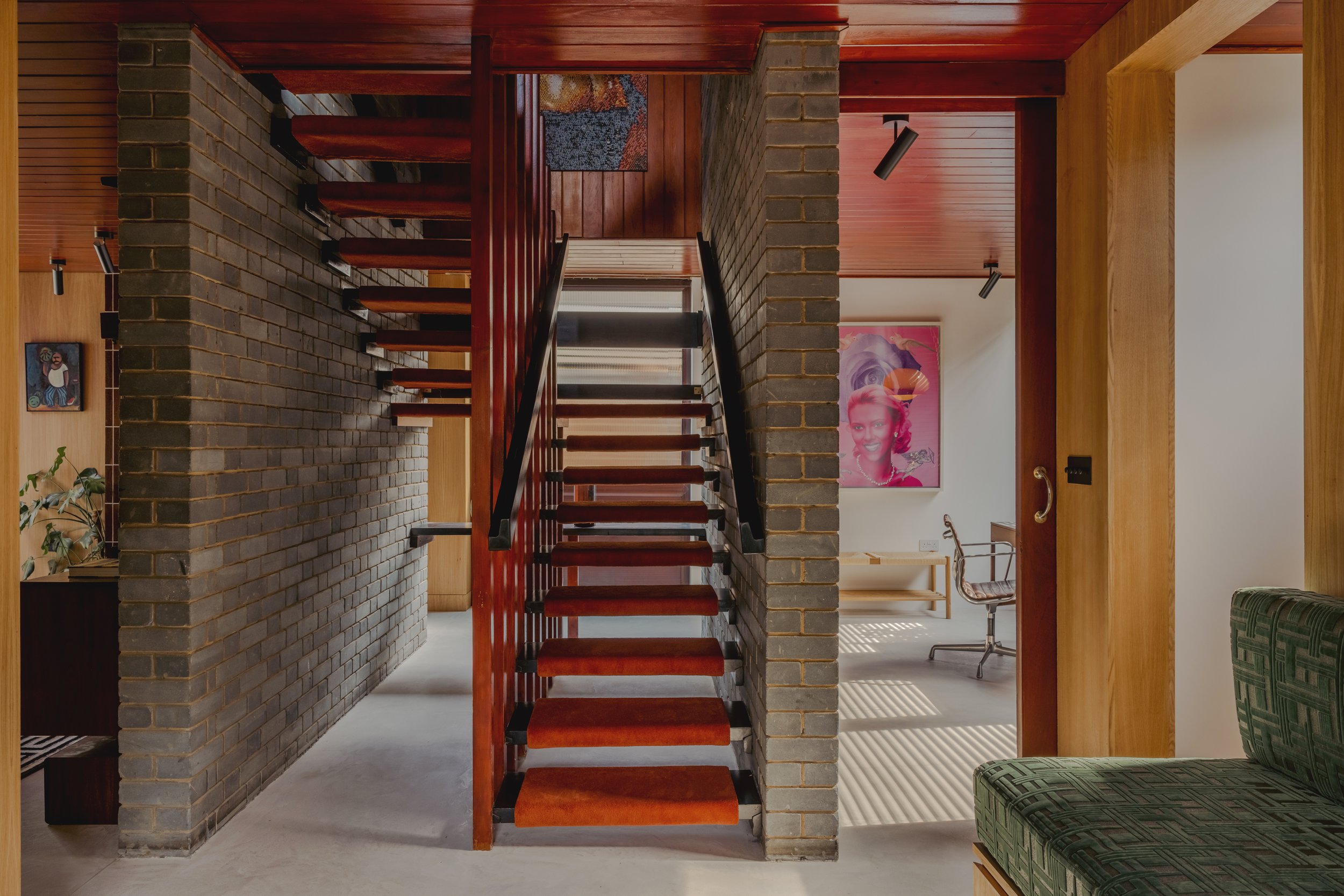 Interior view of a house showing a staircase with wooden steps and black handrails, brick walls, and a room with a desk, office chair, and a Marilyn Monroe poster.