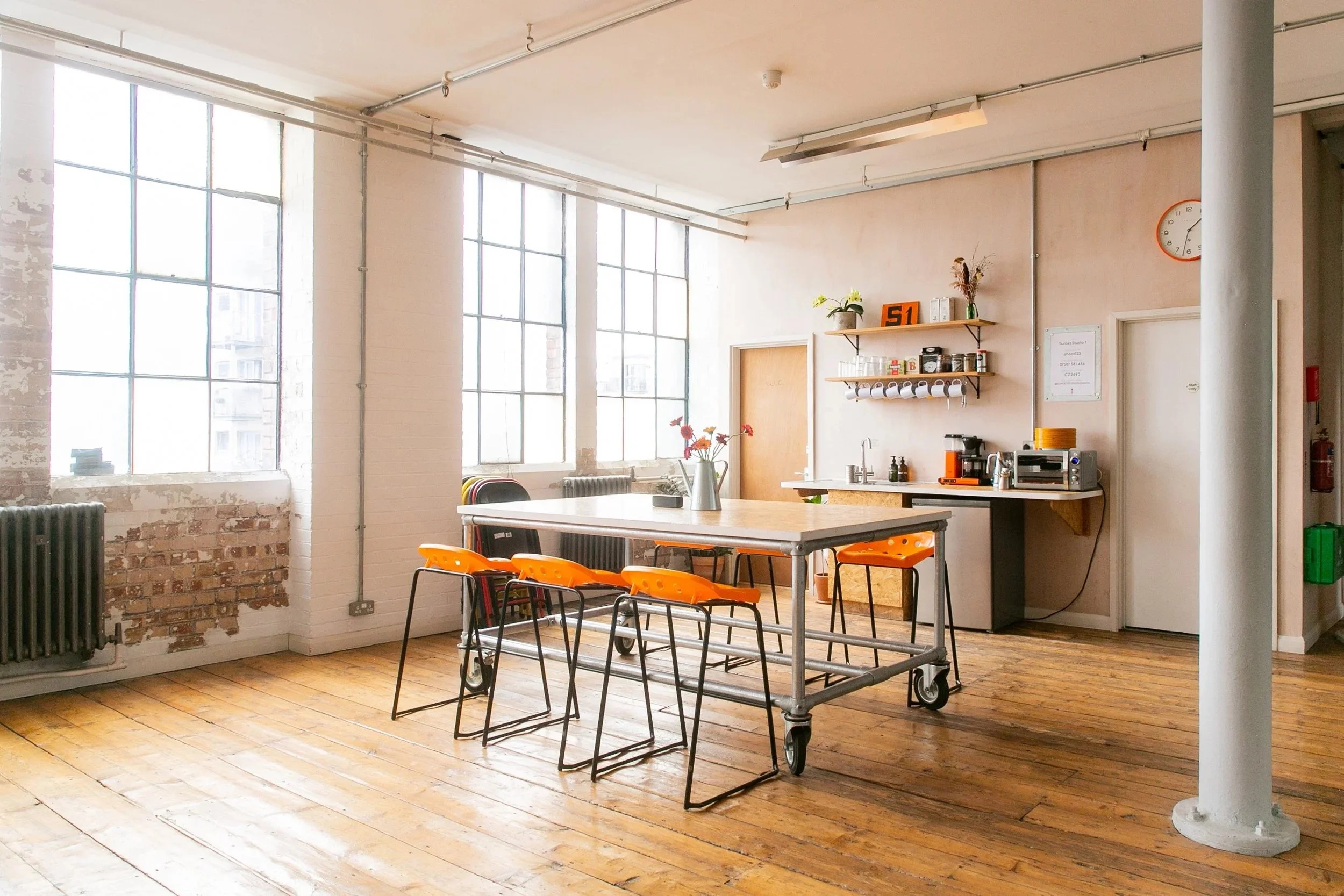 Empty modern kitchen and dining area with large windows, wooden floors, a long table with orange chairs, and a small kitchen setup with shelves and appliances.