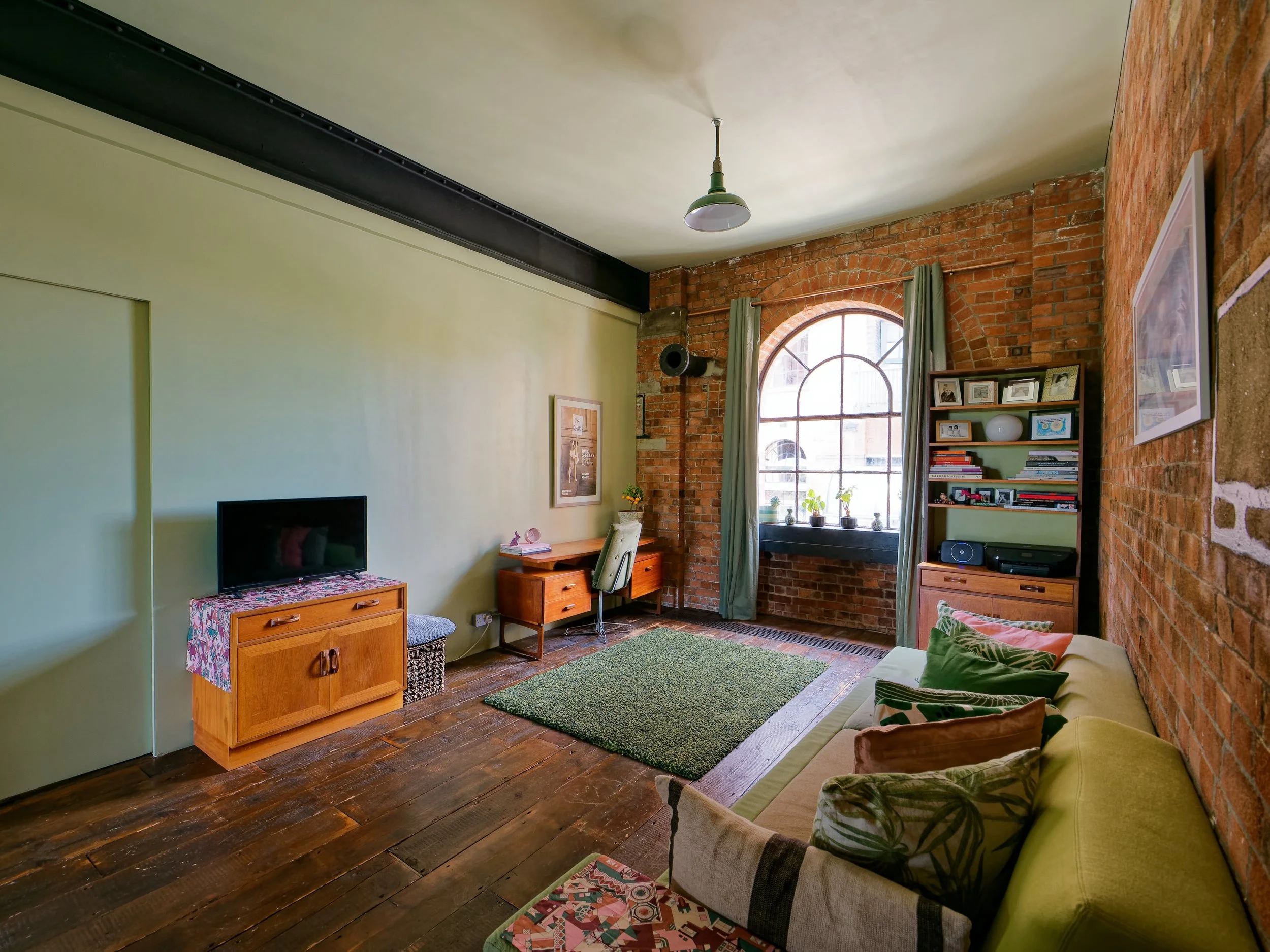 Living room with brick wall, green accent wall, wooden furniture, window with green curtains, sofa with pillows, bookshelf, and a small green rug.