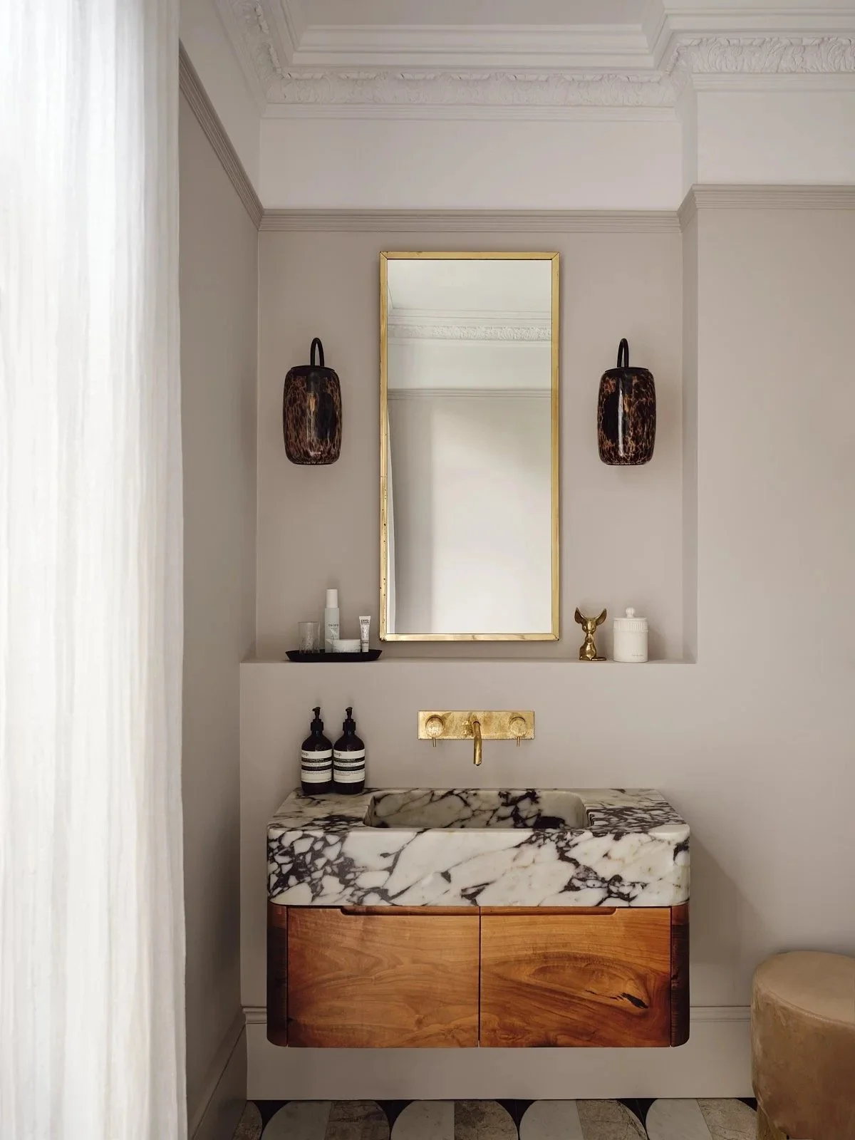 A bathroom vanity with a marble sink, gold faucet, and wooden cabinet, framed mirror, two black and brown wall sconces, and decorative items on shelves.