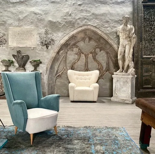 A vintage interior scene with a blue and white armchair, a white beaded armchair, a classical statue on a pedestal, a wrought iron door, and a mural or decorative wall design.