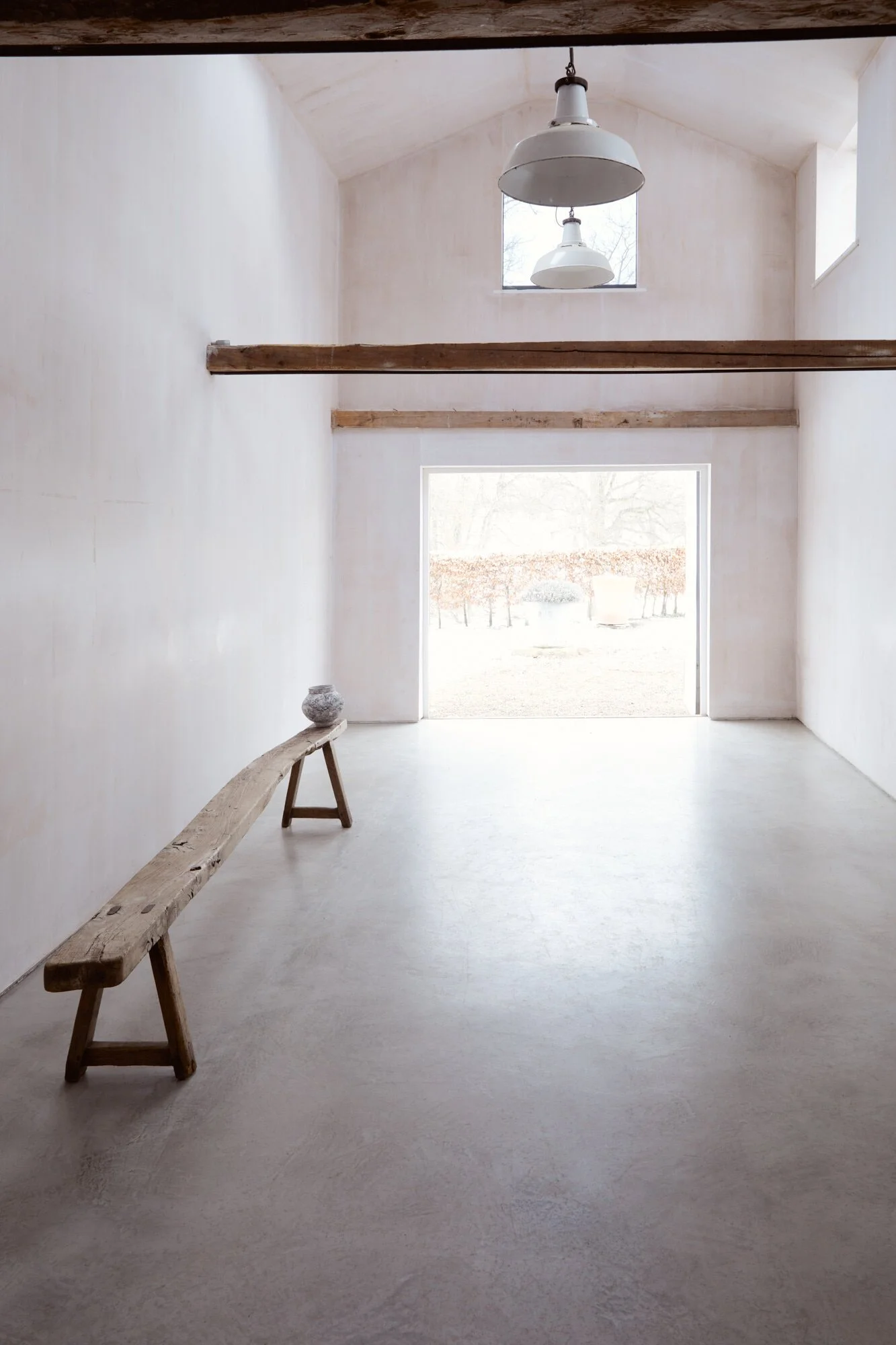 Minimalist room with white walls, a concrete floor, a wooden bench on the left, hanging white pendant lights, exposed wooden beams, and a large open door leading outside with trees and bushes visible.