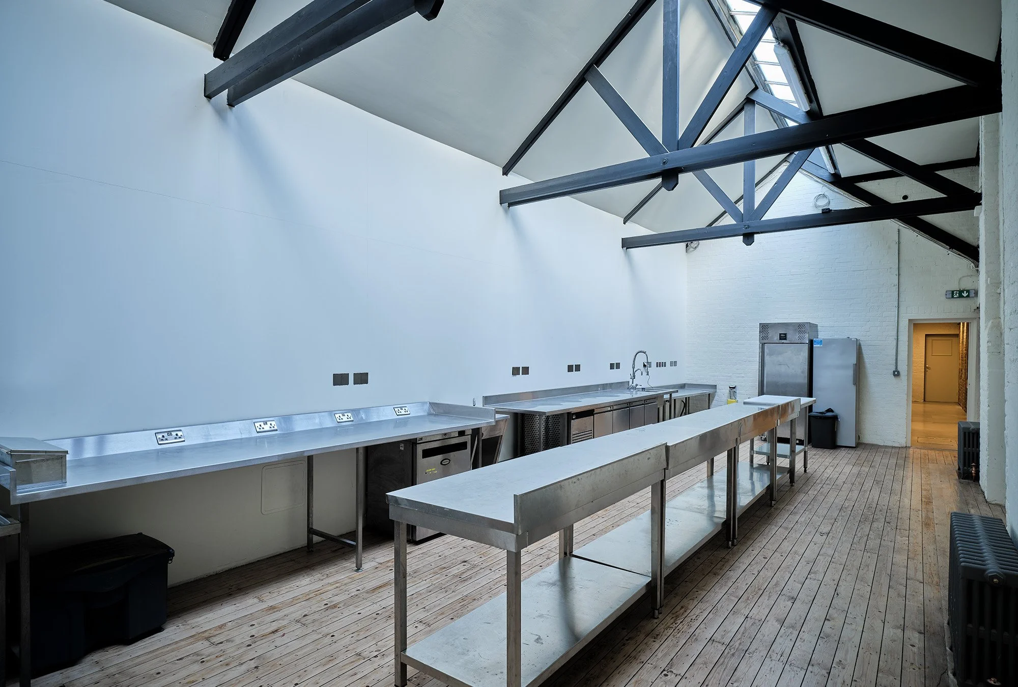 Empty commercial kitchen with stainless steel counters, refrigerator, and a sink under a glass ceiling with exposed beams.
