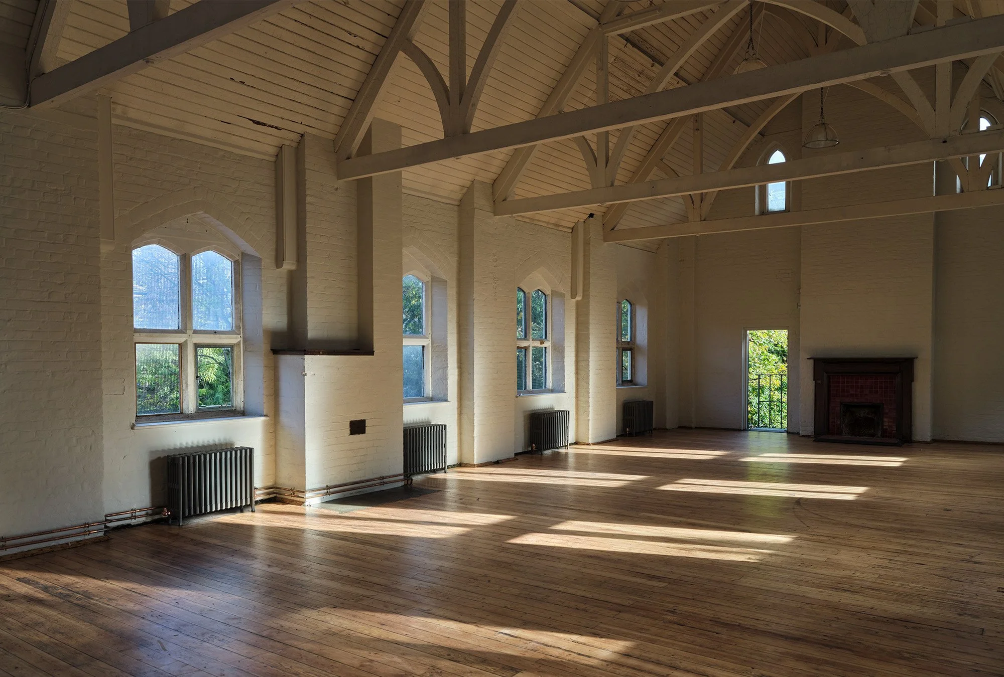 Empty room with wooden floors, white brick walls, large windows, a fireplace, and a vaulted ceiling with wooden beams.