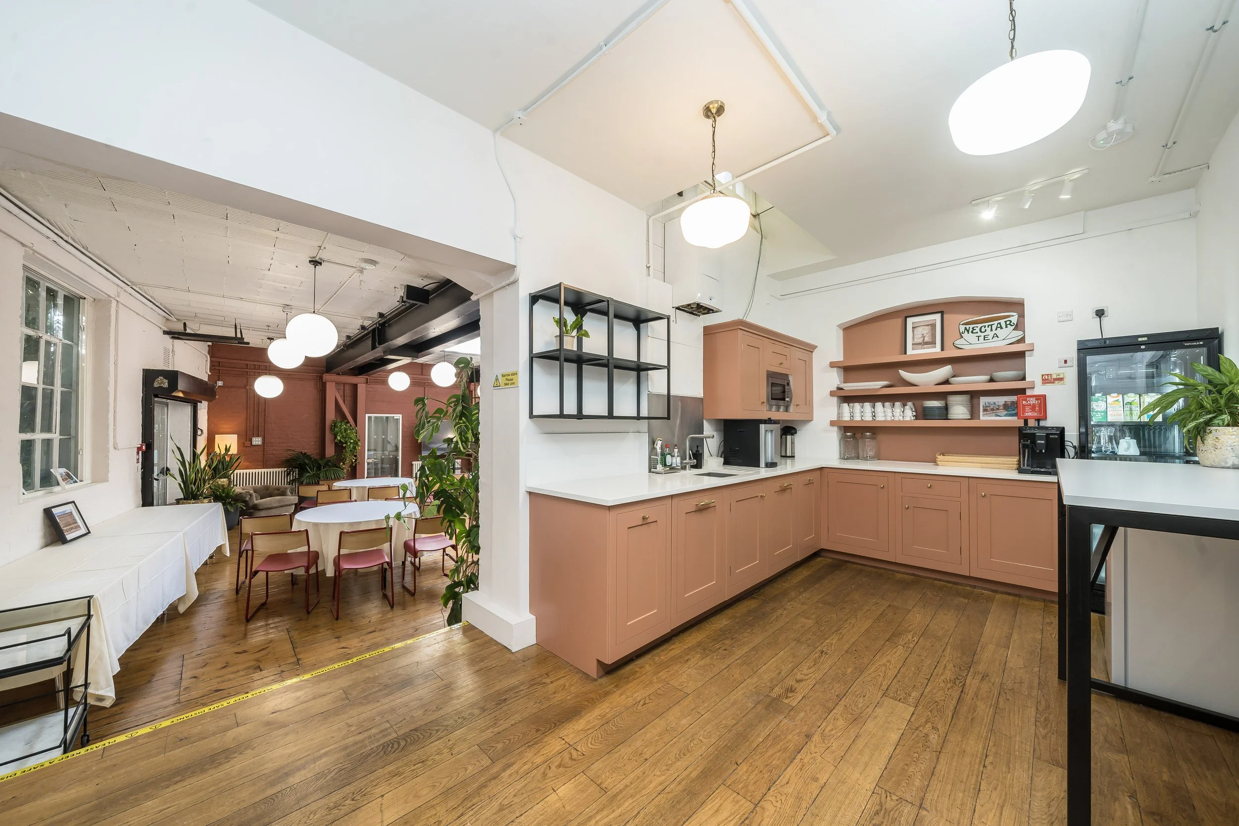 Interior view of a cozy cafe with a kitchen area featuring pinkish cabinets, open shelves with bowls and plates, coffee station, and a small sink. The decor includes potted plants, wooden flooring, and a dining area with round tables, chairs, and lar