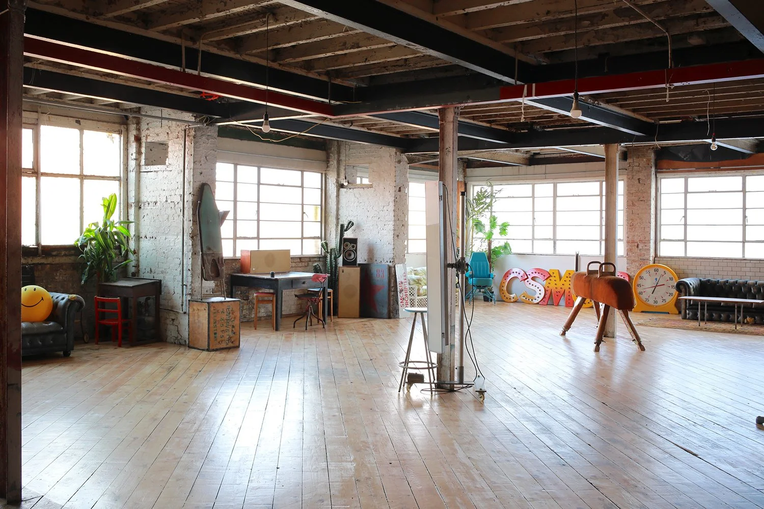 A spacious, well-lit industrial style room with large windows, wooden flooring, exposed brick walls, and a variety of vintage and modern furniture and decor, including plants, a black leather couch, a yellow smiley face pillow, colorful letters spell