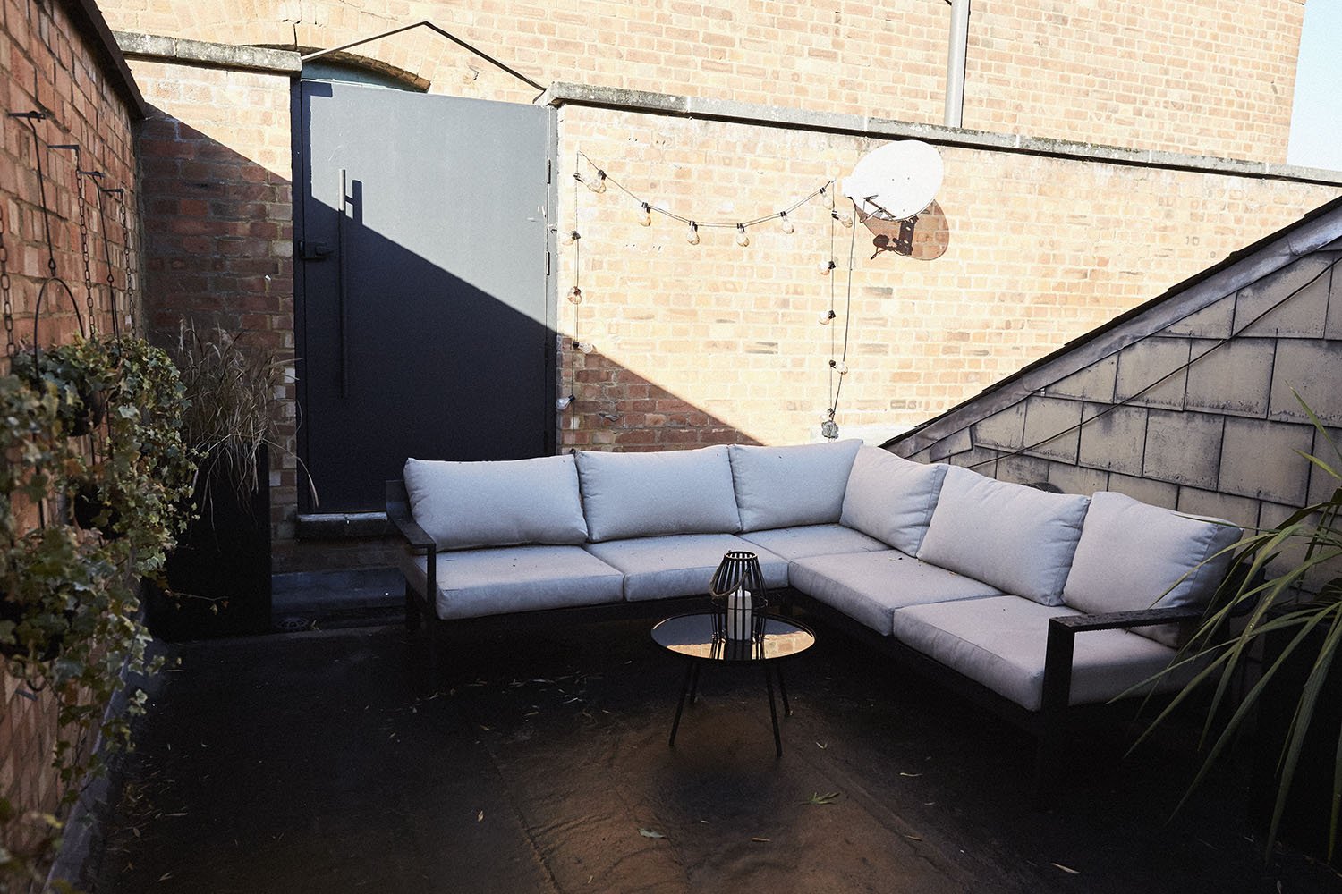 Outdoor patio with an L-shaped white cushioned sofa, a small black metal side table with a decorative lantern, brick walls, string lights, and a satellite dish on the brick wall, with a closed black door.