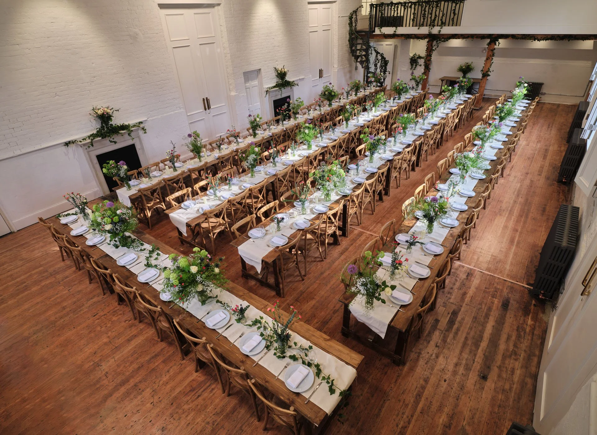 Long banquet tables arranged in a U-shape with white tablecloths and floral centerpieces in a spacious, white-walled room with wooden floors and a staircase.