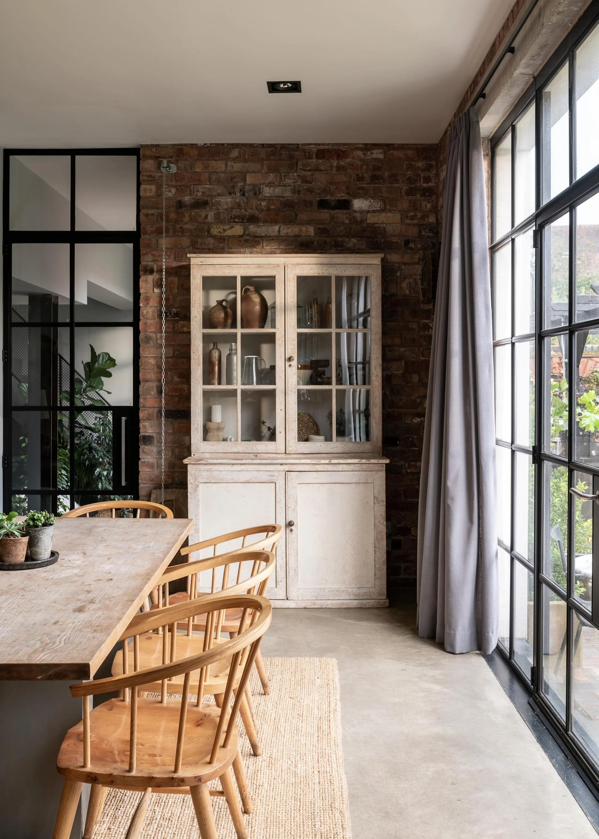 Modern dining area with a wooden table, chairs, and a white vintage cabinet against an exposed brick wall, next to large windows with gray curtains