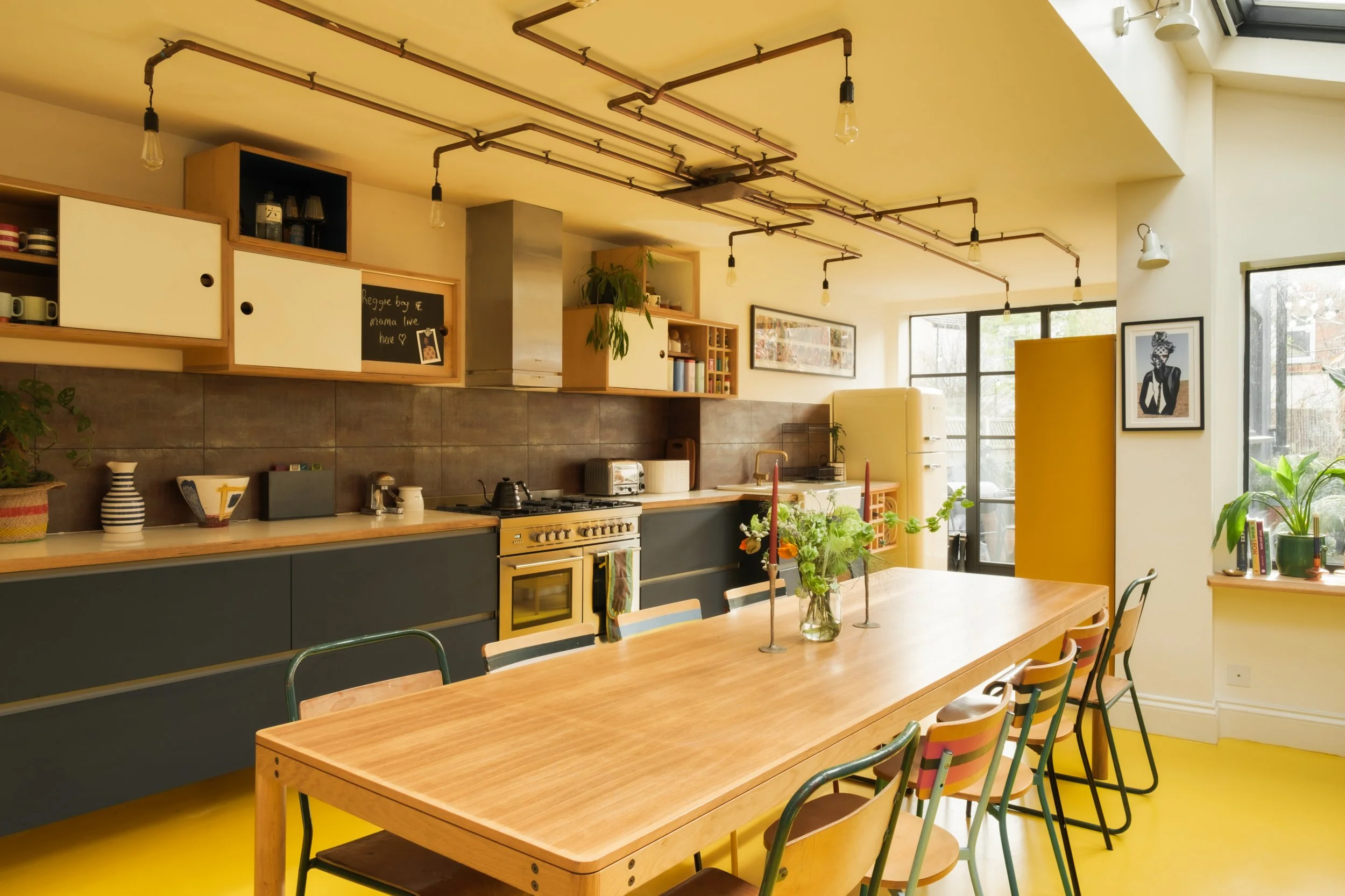 Bright kitchen with wooden dining table, colorful chairs, plants, and natural light from large windows.